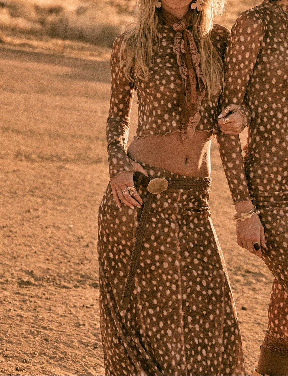 A woman in a desert setting wearing a matching brown and beige polka-dot long-sleeve crop top and skirt set with a wide-brim hat, standing next to another person partially visible on the right.
