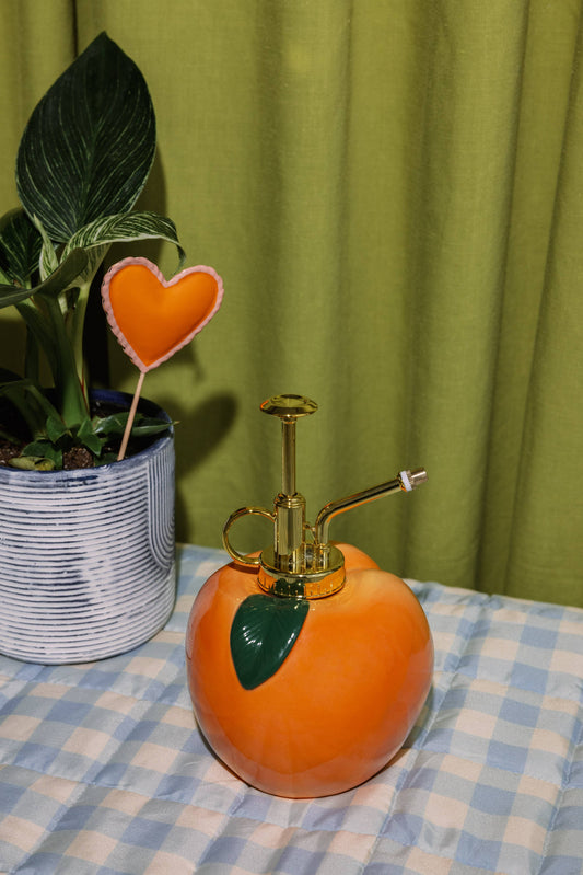 Orange spray bottle with gold nozzle on a checkered tablecloth, next to a potted plant against a green curtain.