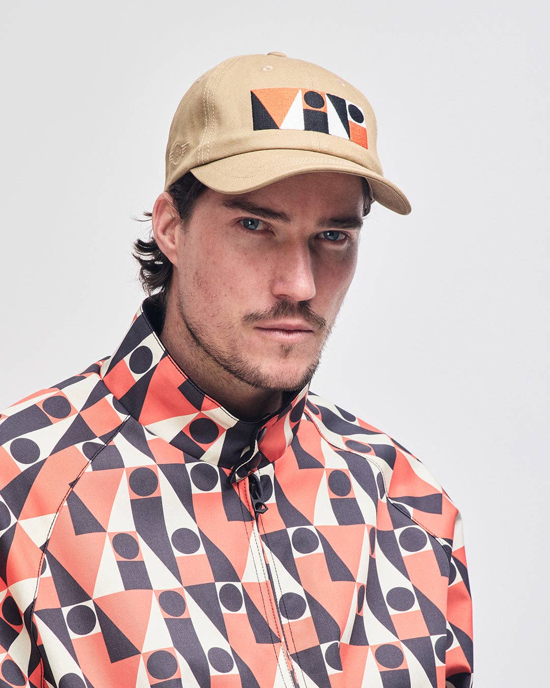 Man wearing a beige cap with geometric design and a patterned shirt on a light gray background