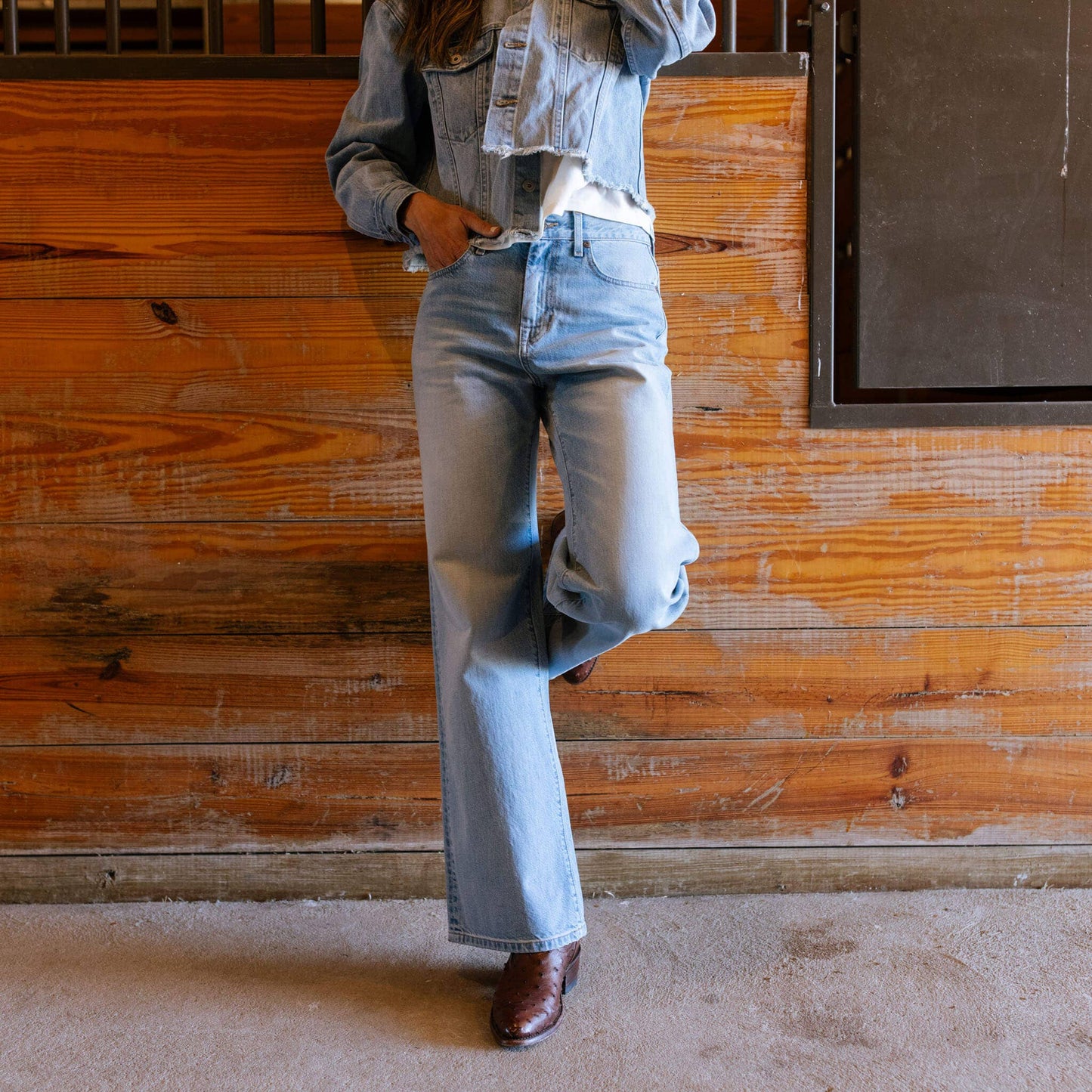 Person wearing light blue jeans and a denim jacket against a wooden wall.