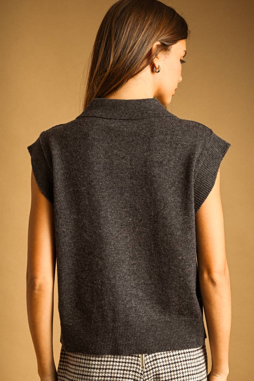 A woman shown from the back wearing a dark charcoal gray sleeveless knit sweater vest over a black and white checkered top, set against a solid warm tan background.
