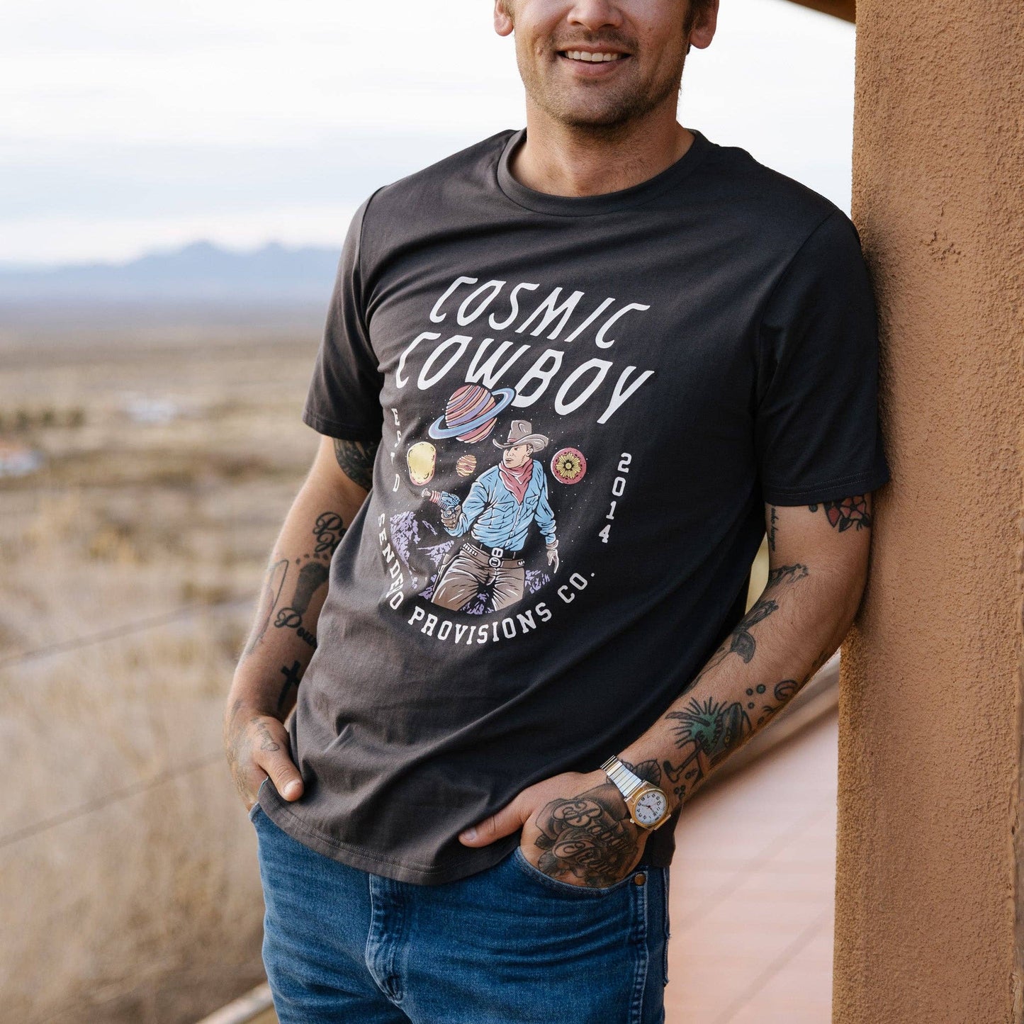 Man wearing a black t-shirt with 'Cosmic Cowboy' design, standing against a desert backdrop.