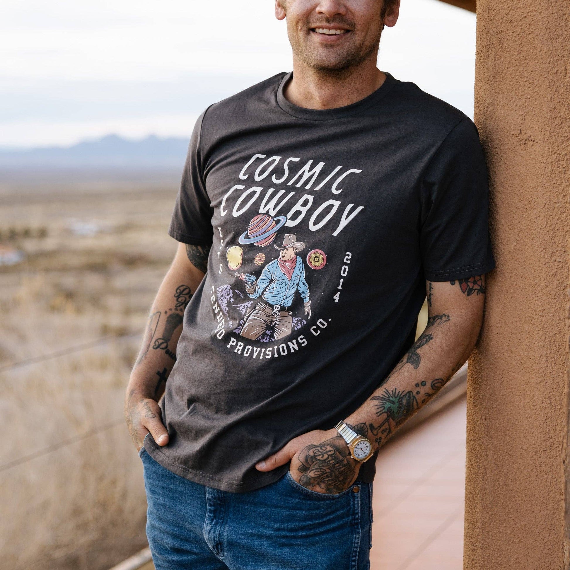 Man wearing a black t-shirt with 'Cosmic Cowboy' design, standing against a desert backdrop.