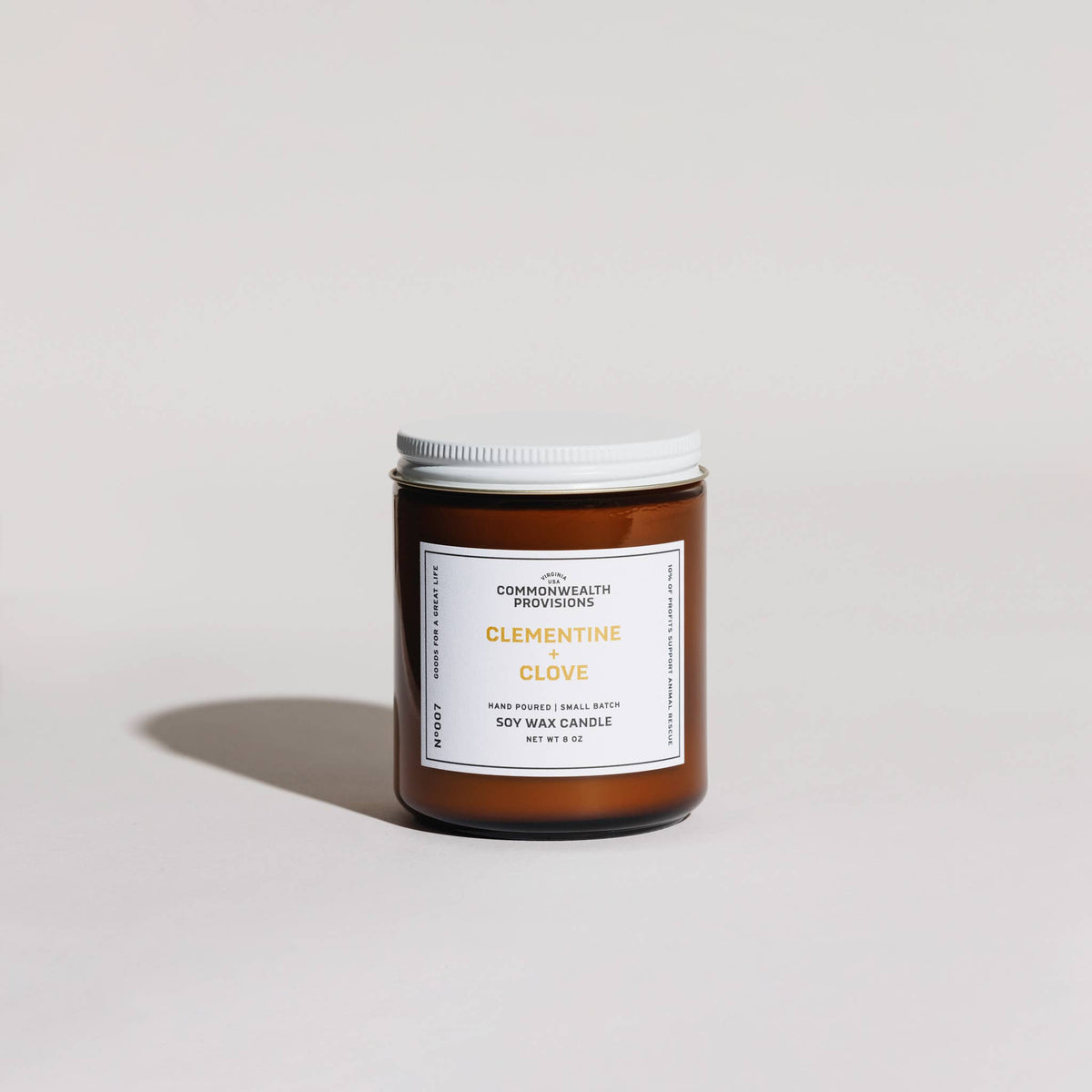 A single amber glass soy wax candle jar with a white lid and a white front label reading 'CLEMENTINE CLOVE' and 'COMMONWEALTH PROVISIONS,' photographed on a clean light gray studio background.