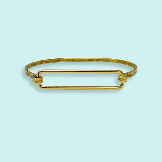 A gold-toned metal bangle bracelet with a textured outer band and a smooth central rectangular bar, shown on a light blue background.