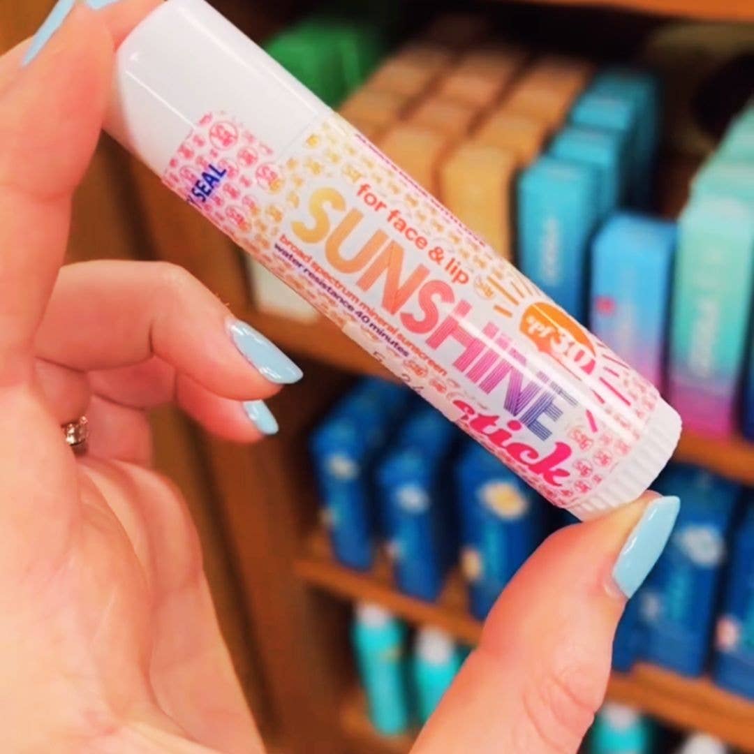 Hand holding a Sunshine stick with a colorful label against a blurred background of shelves.