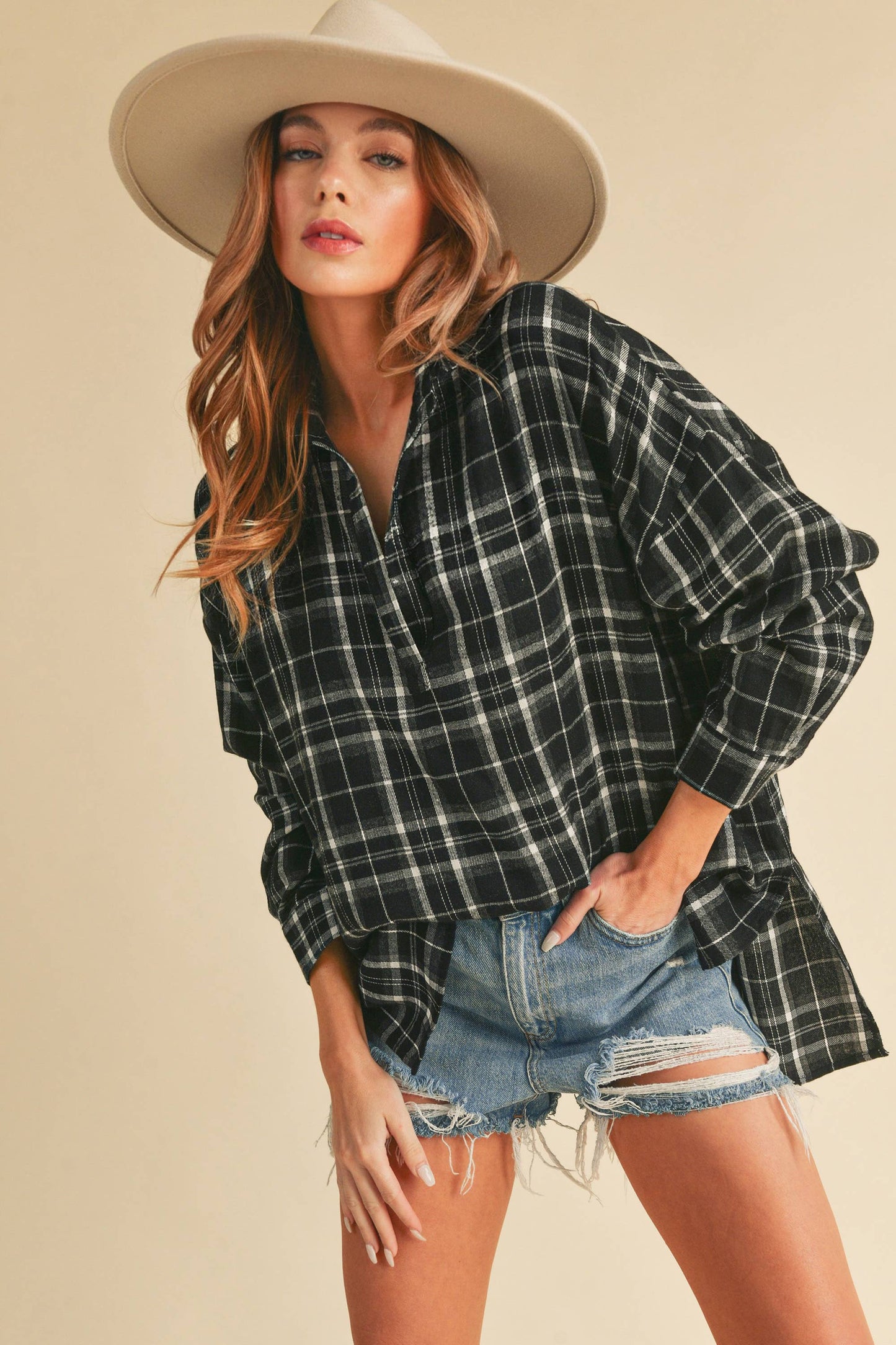 Woman wearing a plaid shirt and denim shorts with a beige hat on a beige background