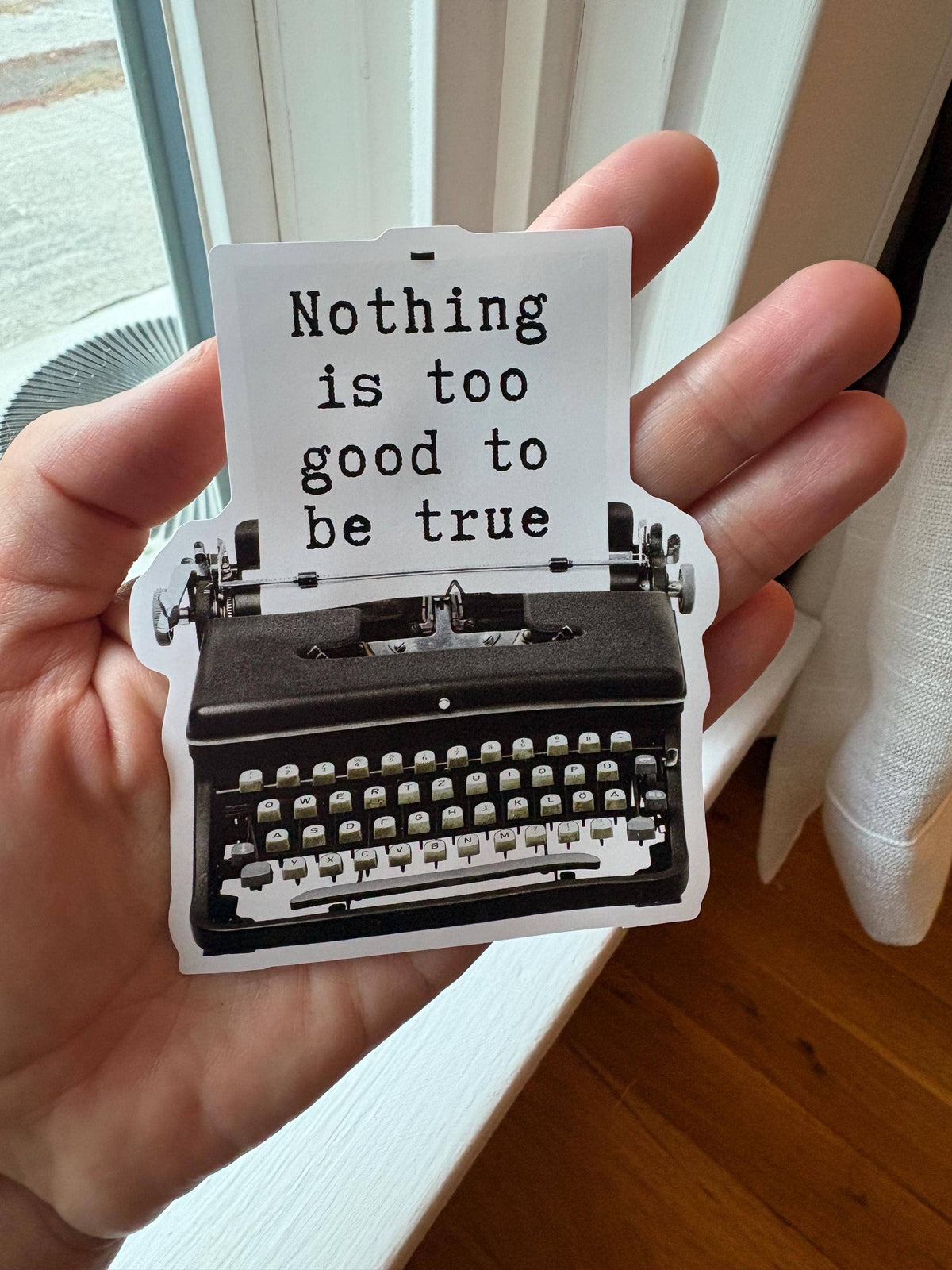 A hand holds a die-cut sticker featuring a black typewriter and the text 'Nothing is too good to be true' in black lettering on a white sheet, with a real indoor background visible.