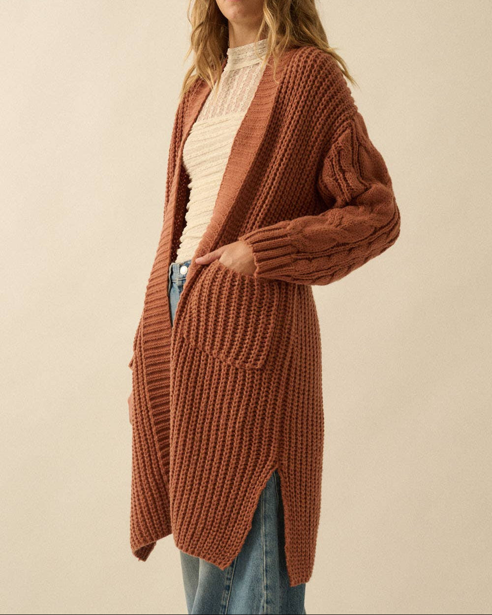 A woman wearing a long, chunky ribbed knit cardigan in a warm rust or terracotta color, layered over a cream ribbed top and blue jeans, against a plain beige studio background.