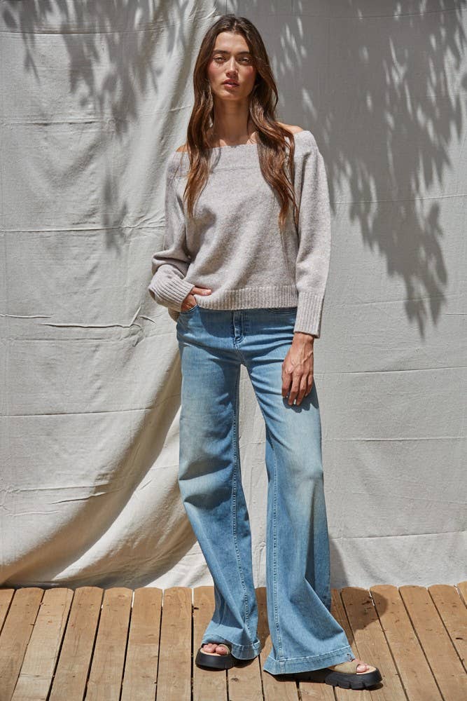 Woman wearing a gray sweater and light blue jeans standing against a plain wall.