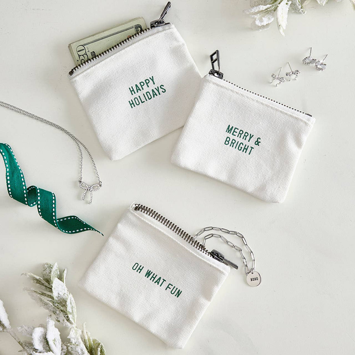 Three small white canvas zip pouches with green holiday text are arranged on a light surface with jewelry and ribbon props. The pouches read 'HAPPY HOLIDAYS,' 'MERRY & BRIGHT,' and 'OH WHAT FUN.