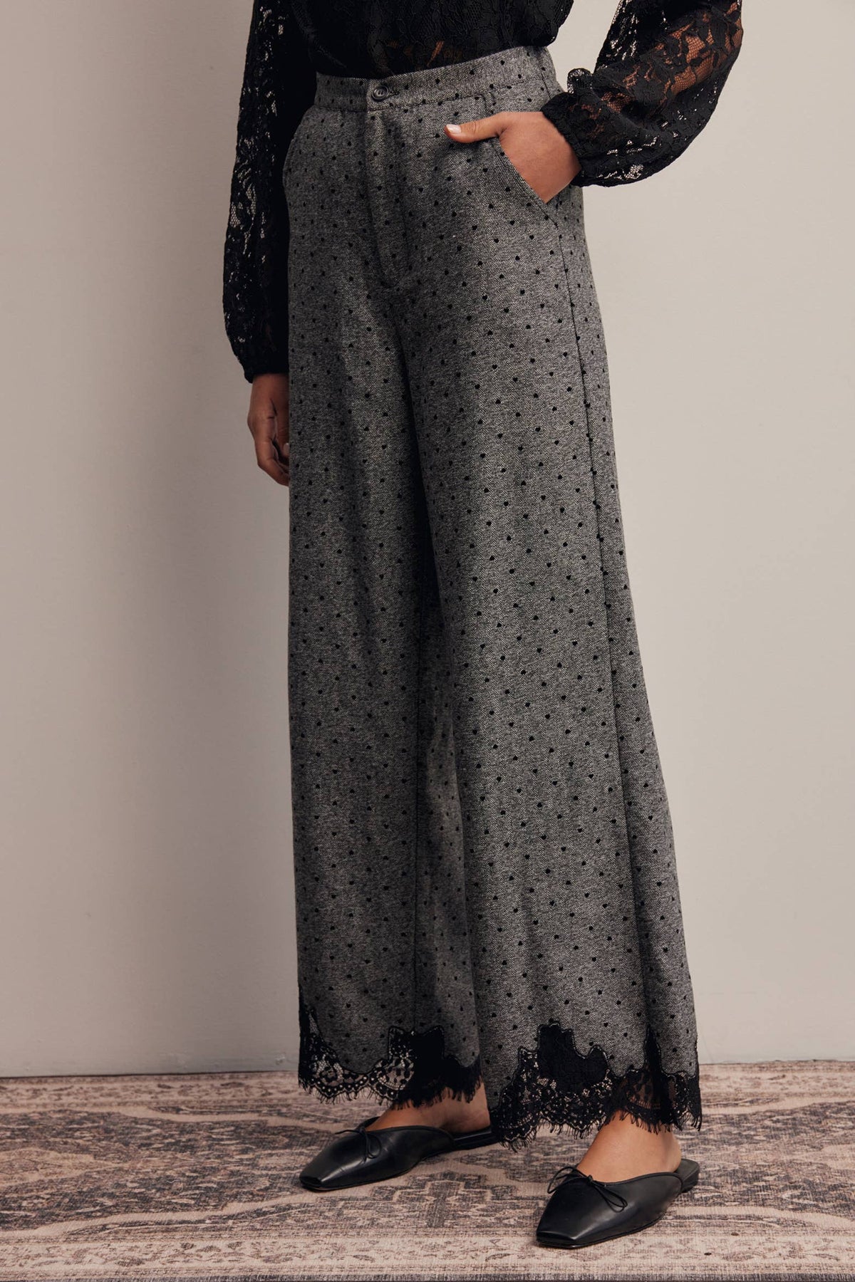 A person wearing high-waisted, wide-leg gray pants with a black floral print and black lace trim at the hem, styled with a black lace long-sleeve top and black pointed flats.