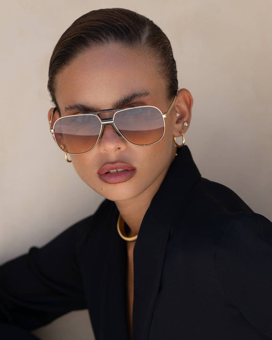Person wearing gold aviator sunglasses and a black outfit against a neutral background