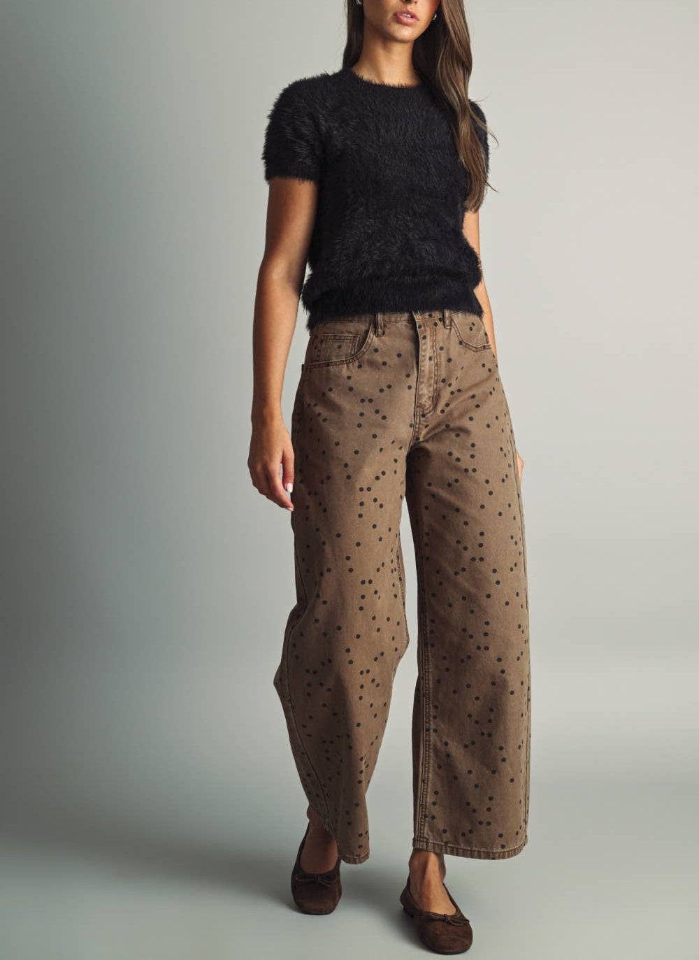 A woman modeling a black fuzzy short-sleeve top and brown wide-leg pants with black polka dots, standing against a neutral gray studio background.