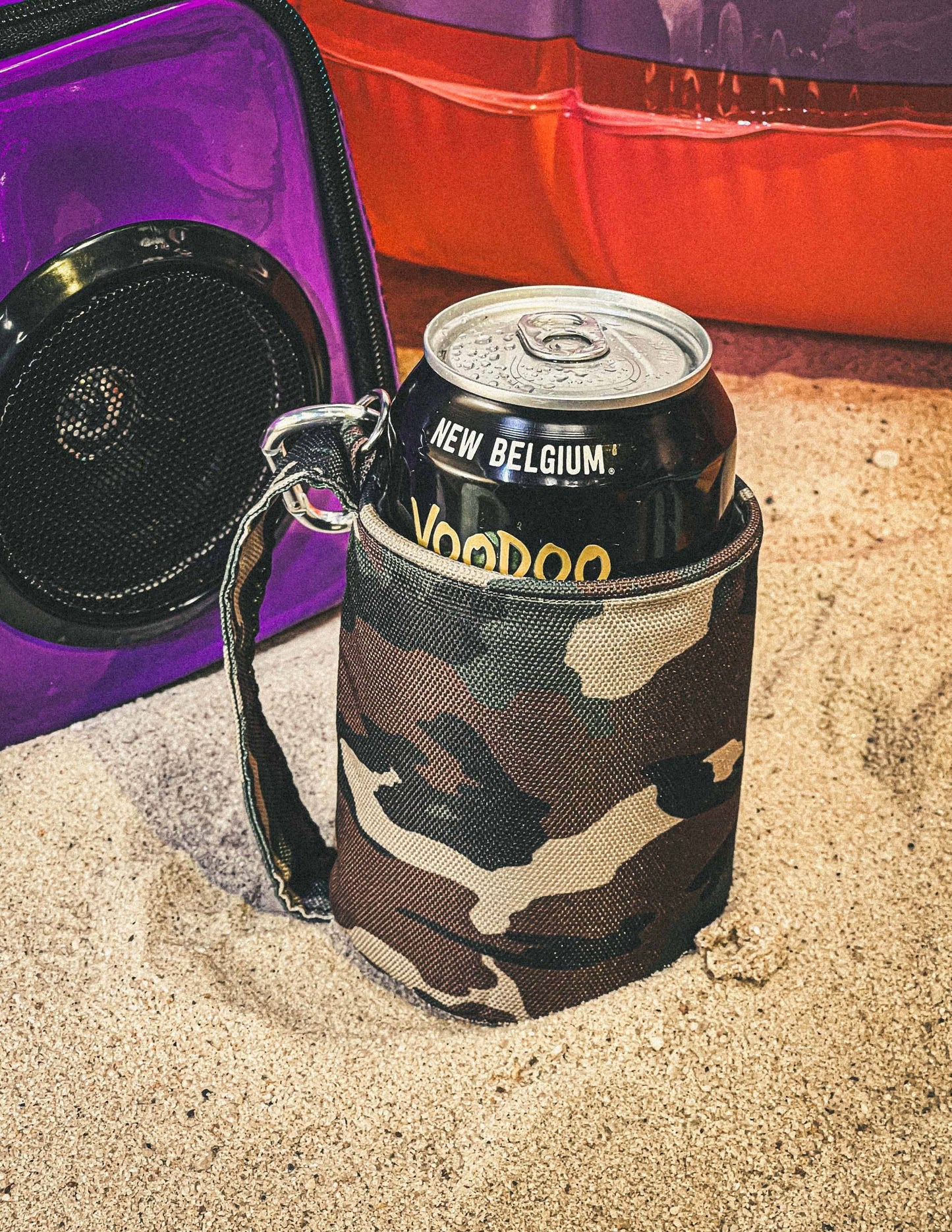 Camouflage can holder with New Belgium Voodoo beer can on sand next to a purple cooler.