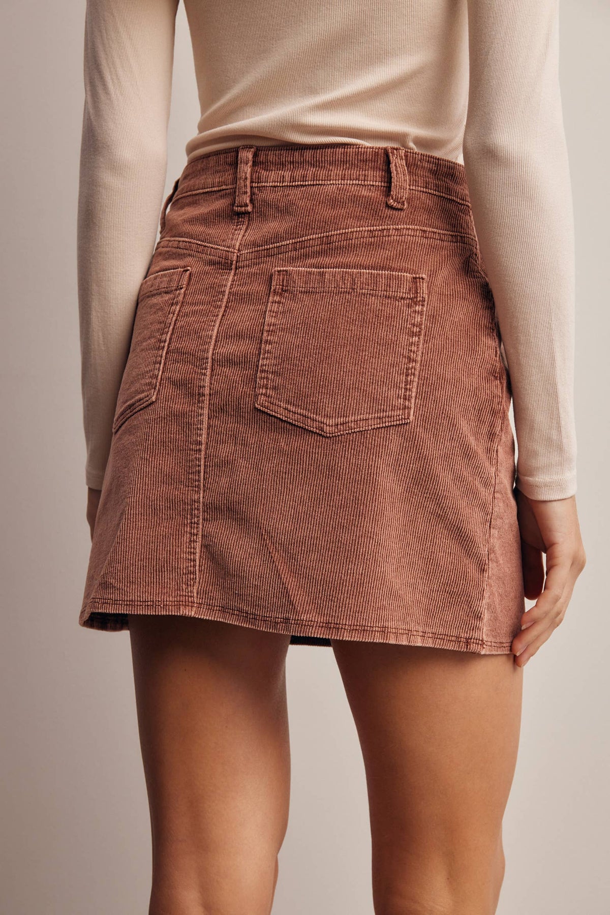 A person wearing a dusty rose corduroy mini skirt with back patch pockets, paired with a beige long-sleeve top, shown from the back against a neutral studio background.