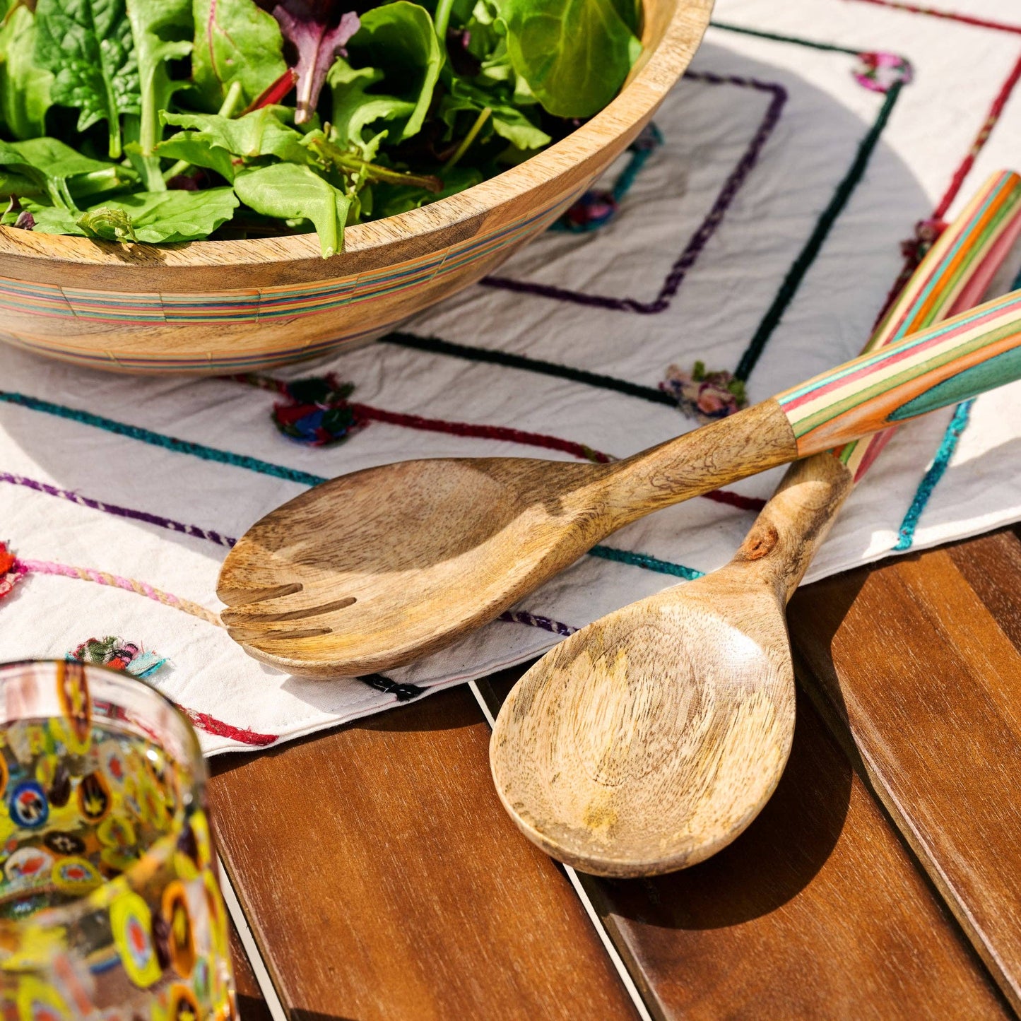 Wooden salad bowl with salad, wooden spoons, and a colorful glass on a patterned tablecloth.