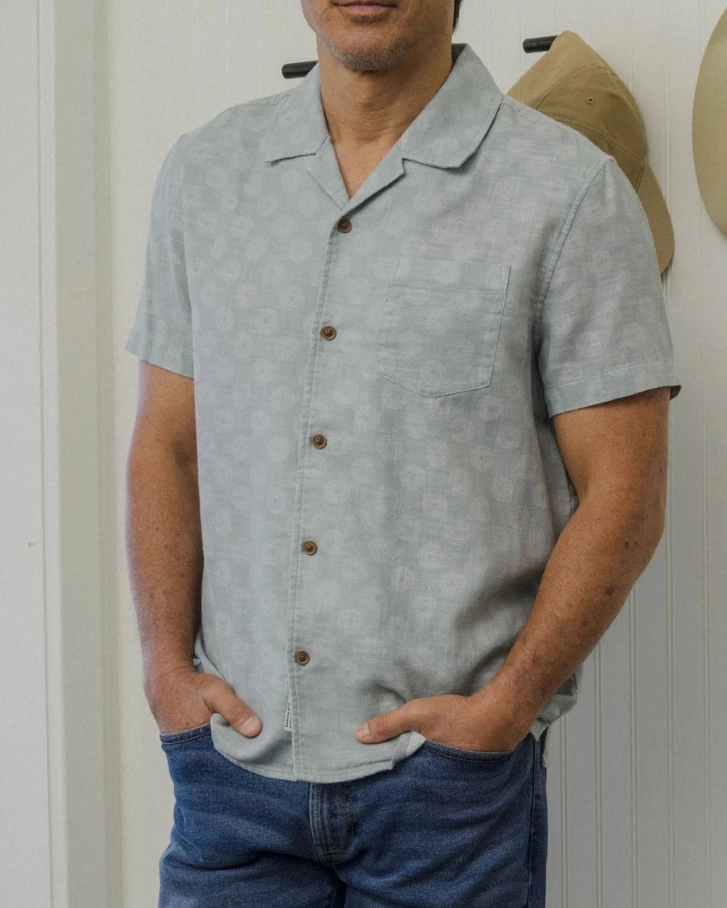Man wearing a light gray shirt and blue jeans standing in a room with hats on the wall.