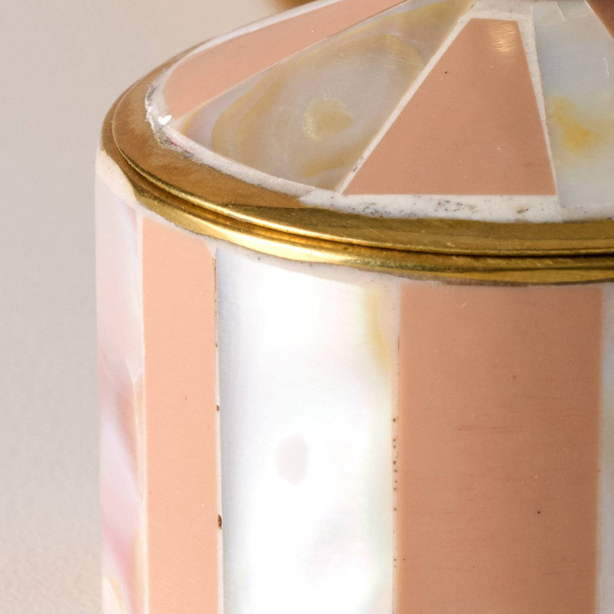 Close-up of a cylindrical decorative container or lamp base featuring vertical stripes of iridescent white, pale pink, and peach, accented by a polished gold metal rim and a geometric patterned top.