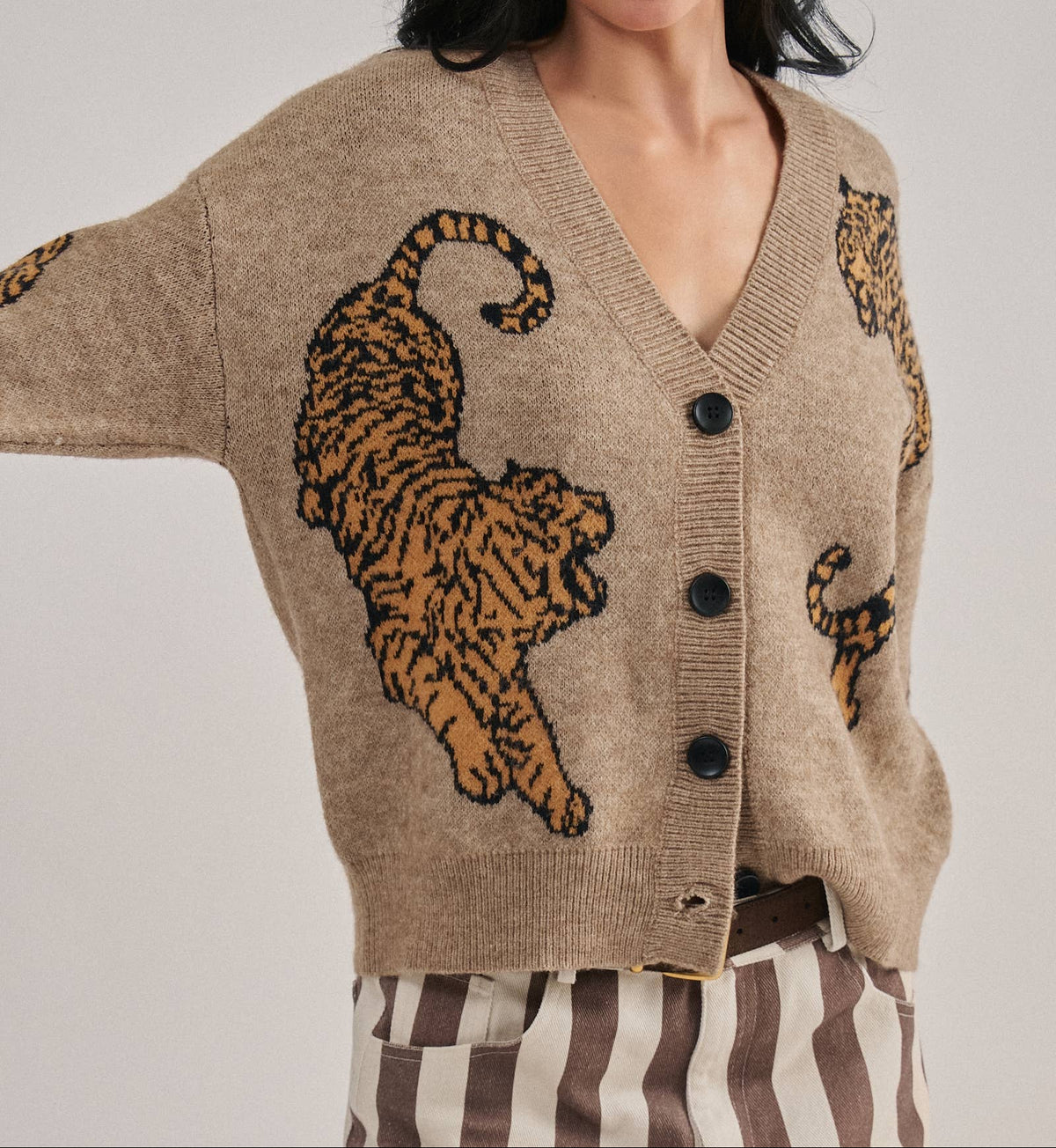 A woman wearing a beige knit cardigan with large tiger motifs in orange and black, paired with brown and white striped pants, against a light gray studio background.