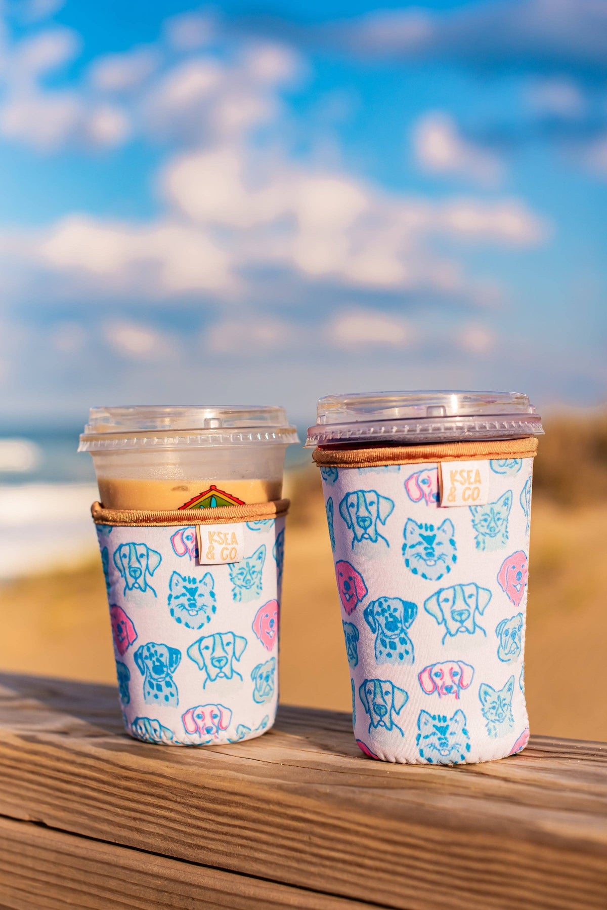Two coffee cups with colorful elephant patterns on a wooden surface with a beach background.