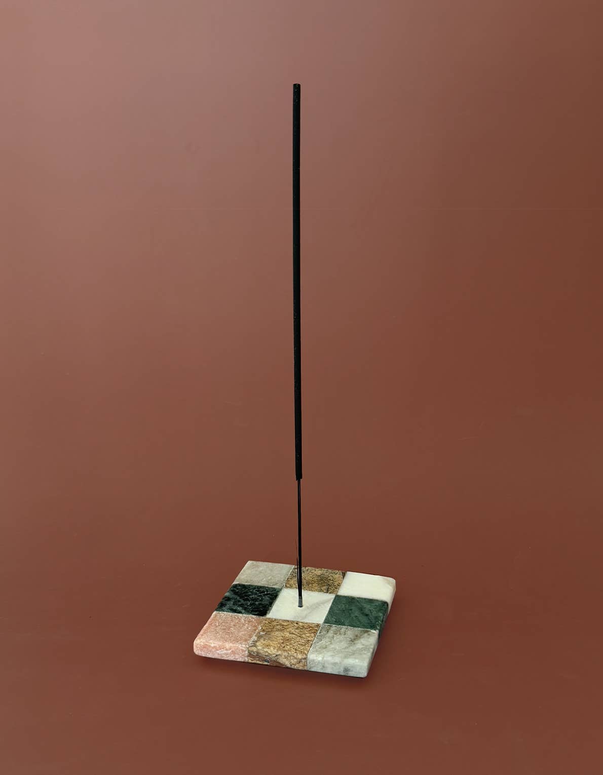 Incense holder with a square, multicolored base and a tall, thin black rod on a brown background