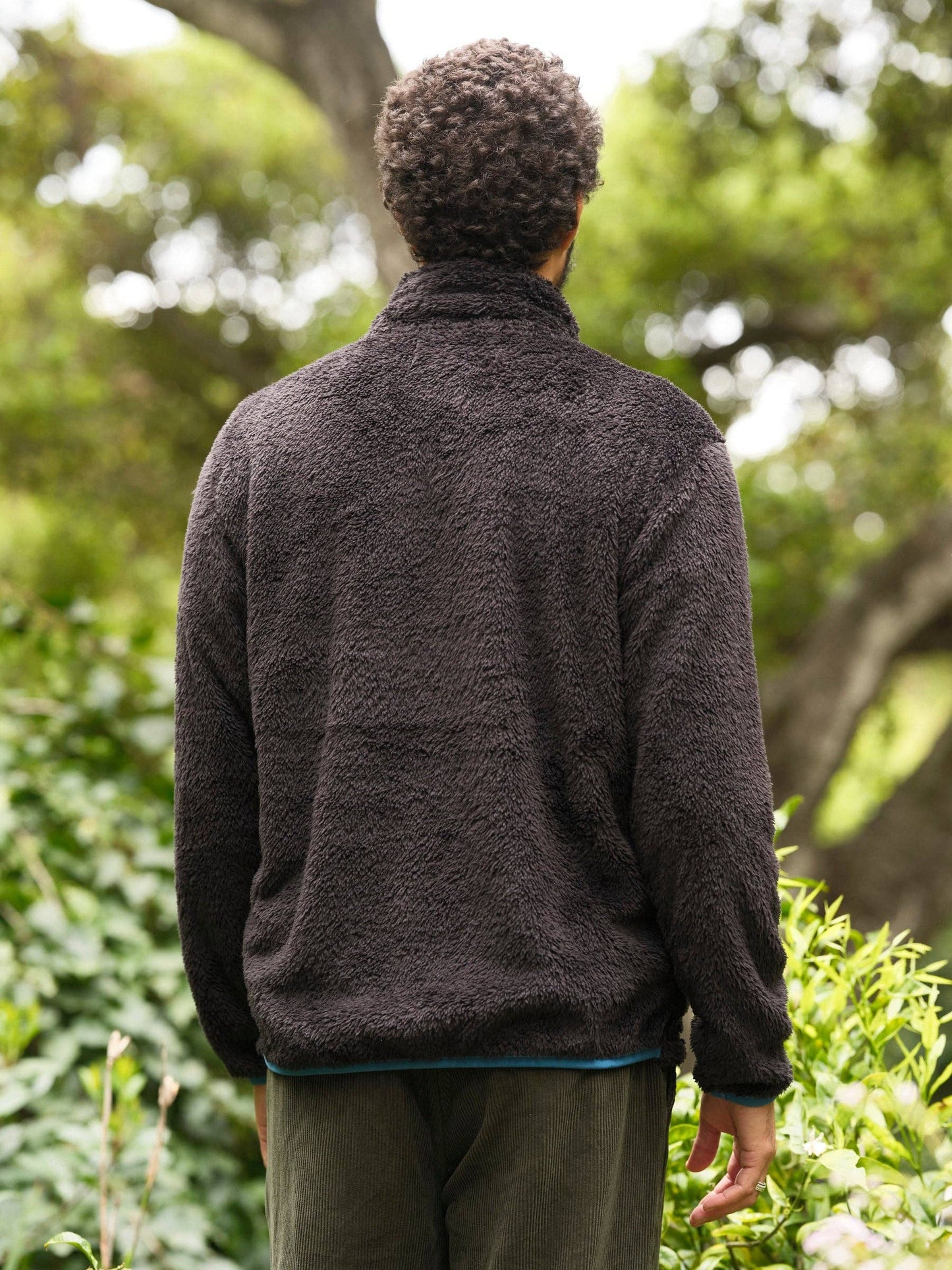 Person wearing a dark jacket standing in a natural setting with trees and greenery.