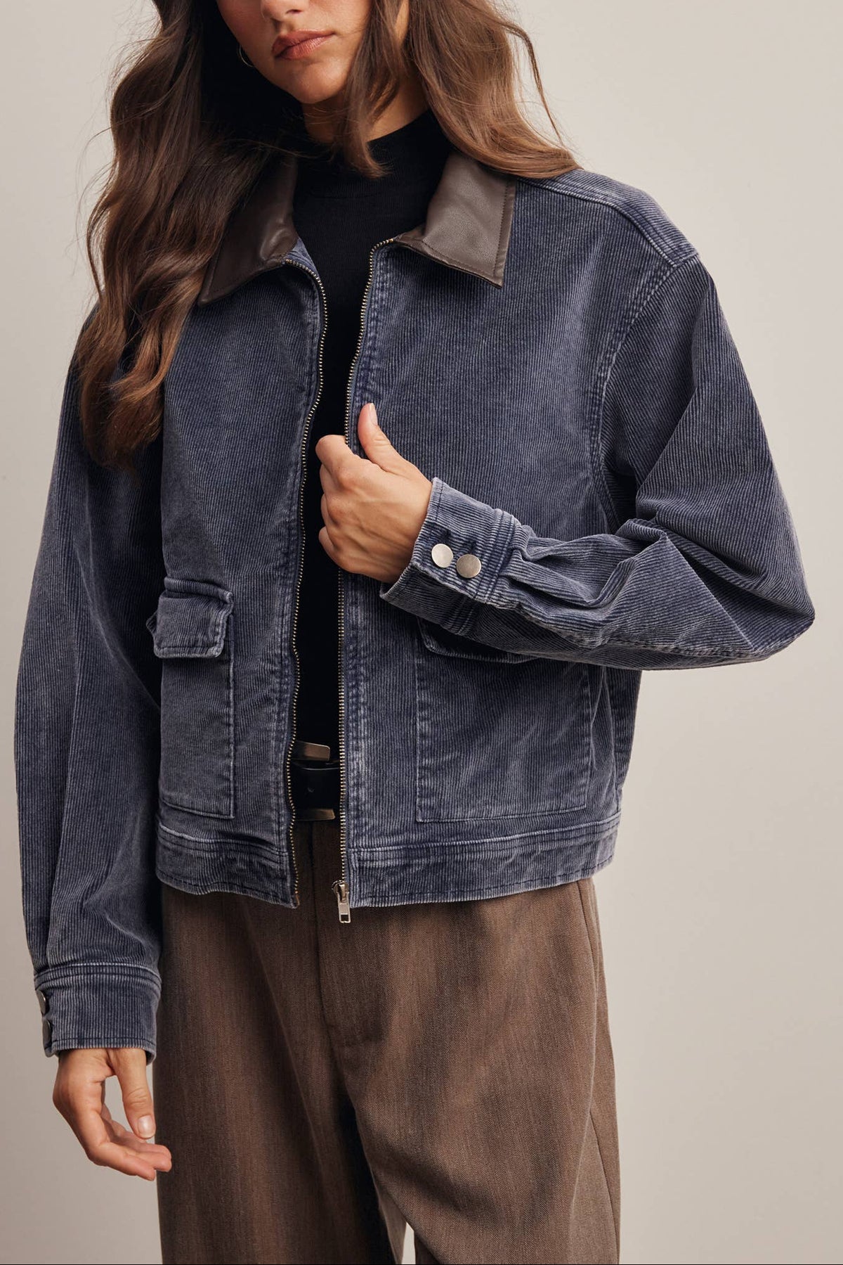 A woman wearing a dark blue washed corduroy jacket with a brown collar, styled over a black top and brown trousers, against a plain beige studio background.