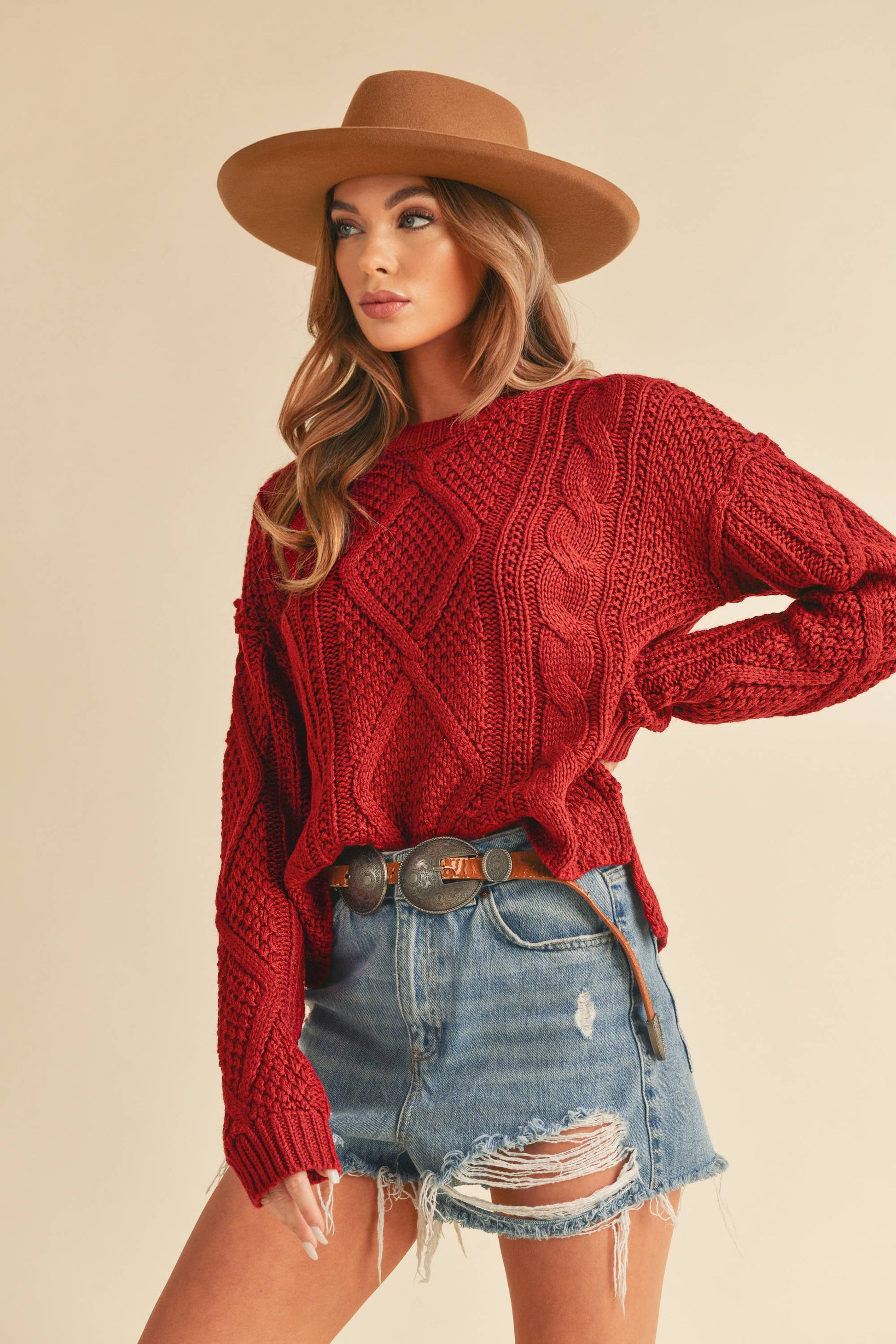 Woman wearing a red knitted sweater, denim shorts, and a brown hat on a beige background