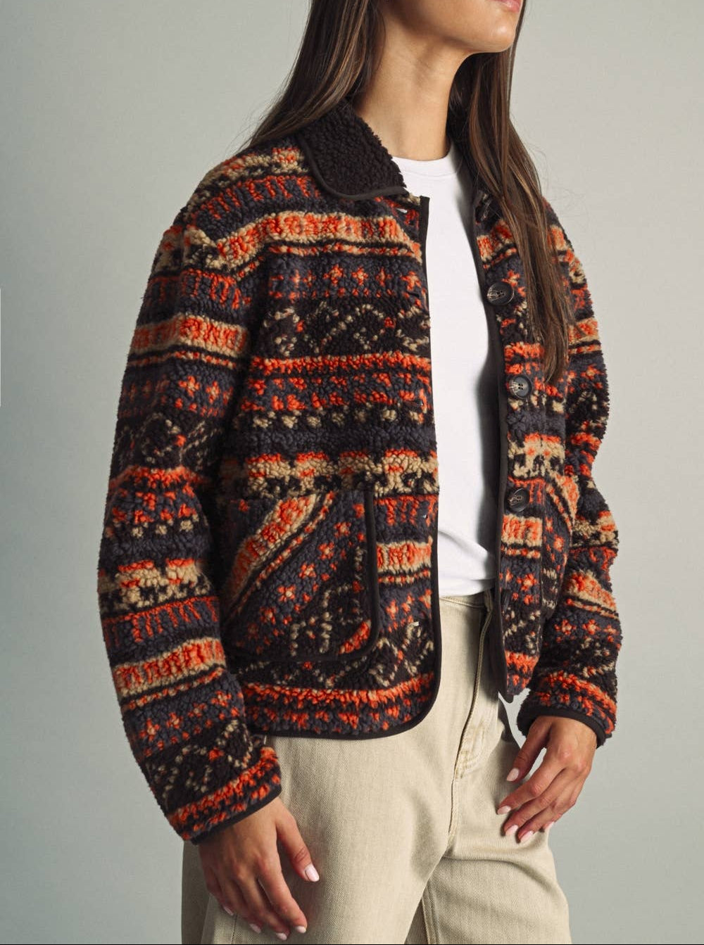 A woman wearing a patterned knit zip-up jacket with a dark brown sherpa collar, styled with a white t-shirt and beige pants against a neutral gray background.