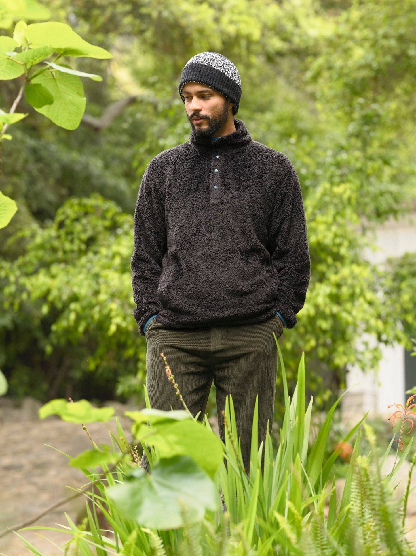 Man wearing a dark sweater and knit hat standing in a garden with greenery.