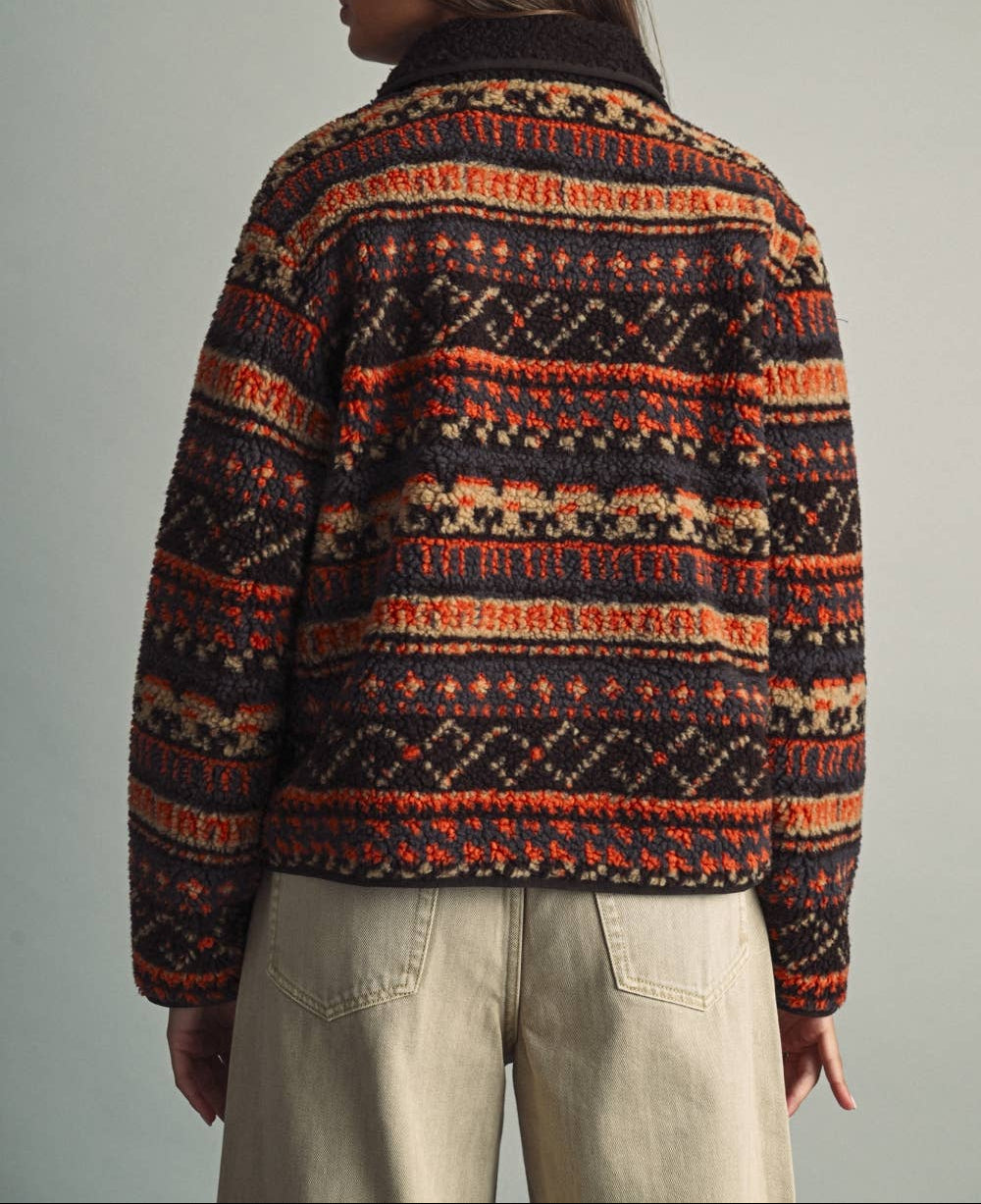 A person shown from the back wearing a dark knit sweater with horizontal Fair Isle-style patterns in orange, cream, and beige, paired with light beige pants, against a plain light gray studio background.