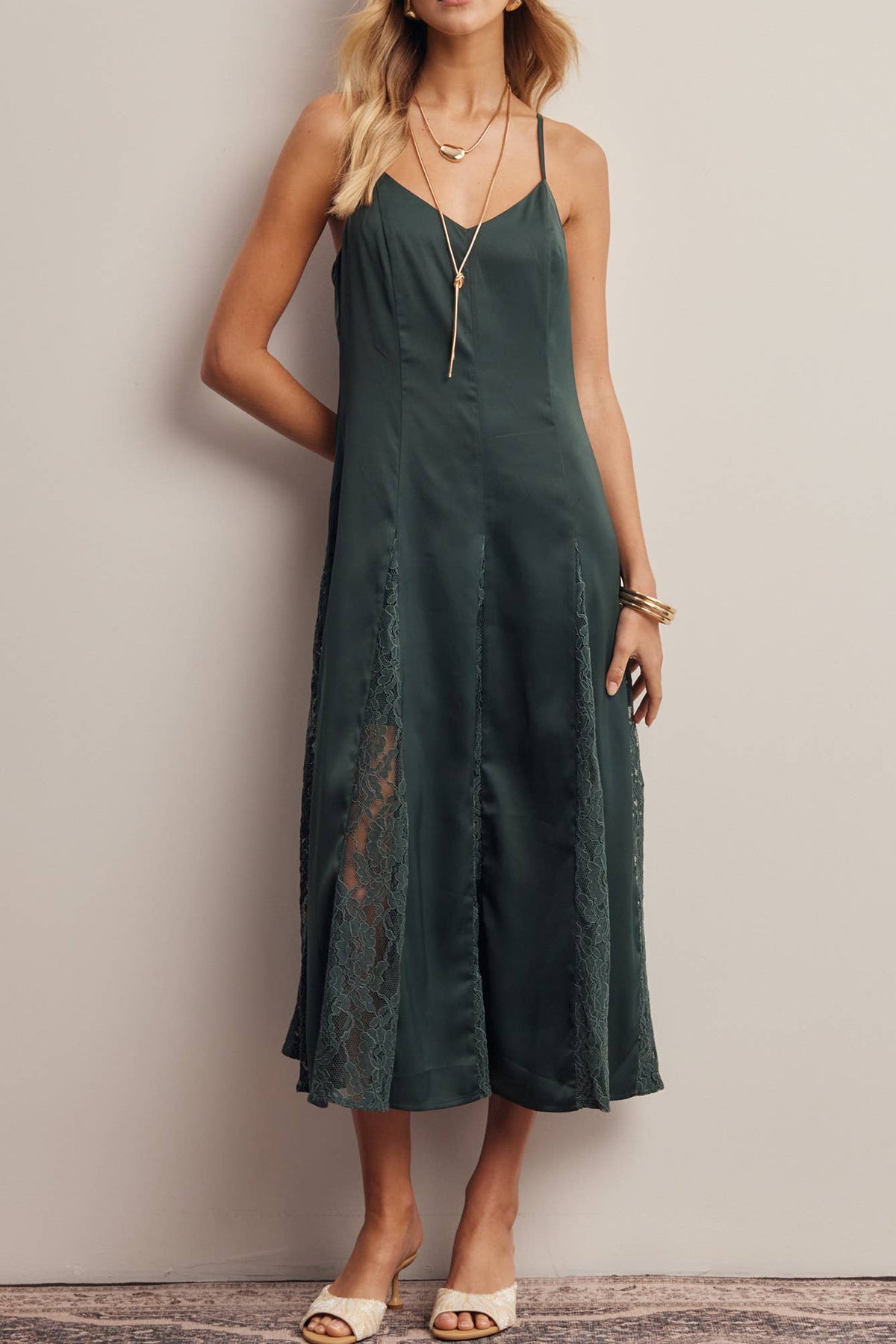 A woman models a dark green sleeveless midi dress with a lace overlay and spaghetti straps, standing against a neutral beige wall.