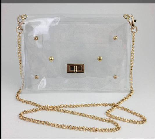 A clear vinyl shoulder bag with gold-tone hardware, including a chain strap, gold studs, and a central metal clasp, shown on a plain light background.