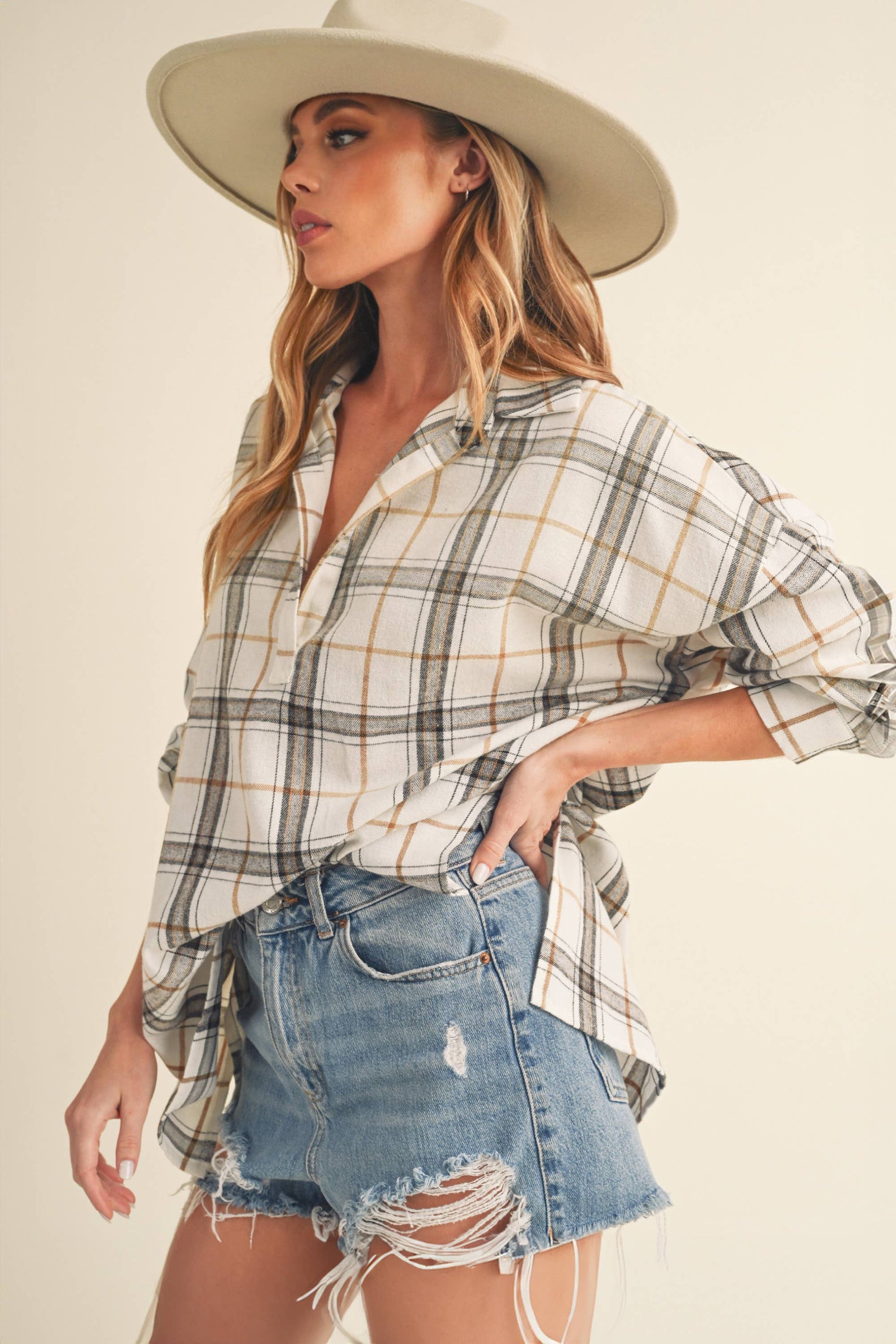 Woman wearing a plaid shirt and denim shorts with a beige hat on a plain background