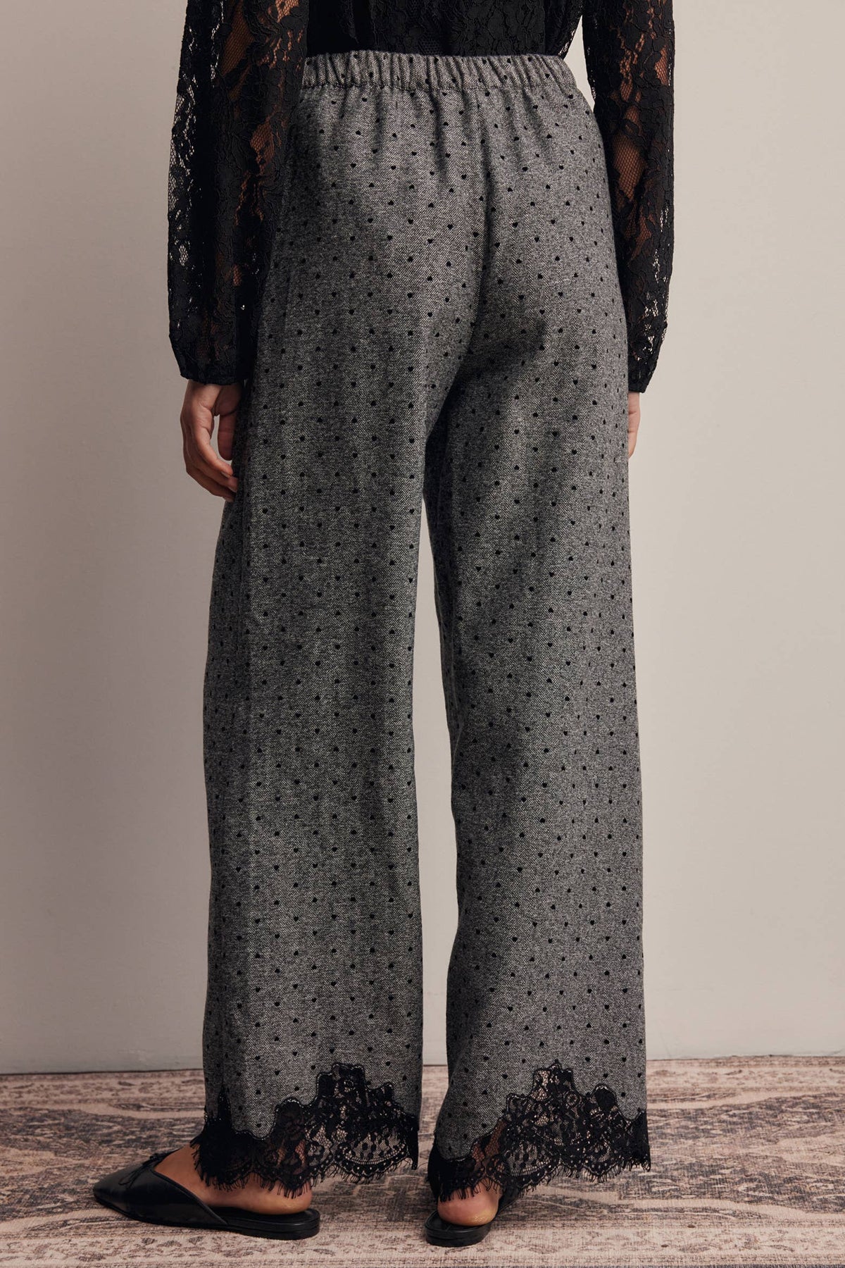 A person shown from the back wearing wide-leg gray pants with a small black dot pattern and black lace trim at the hems, paired with black slip-on shoes against a plain beige wall.