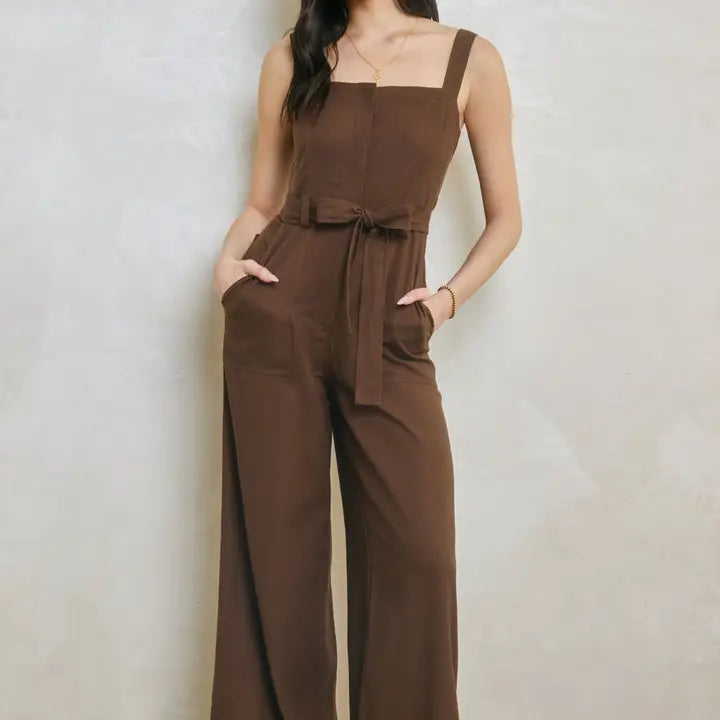 A woman wearing a brown sleeveless jumpsuit with a square neckline, tie waist, and wide legs, standing against a textured light beige wall.