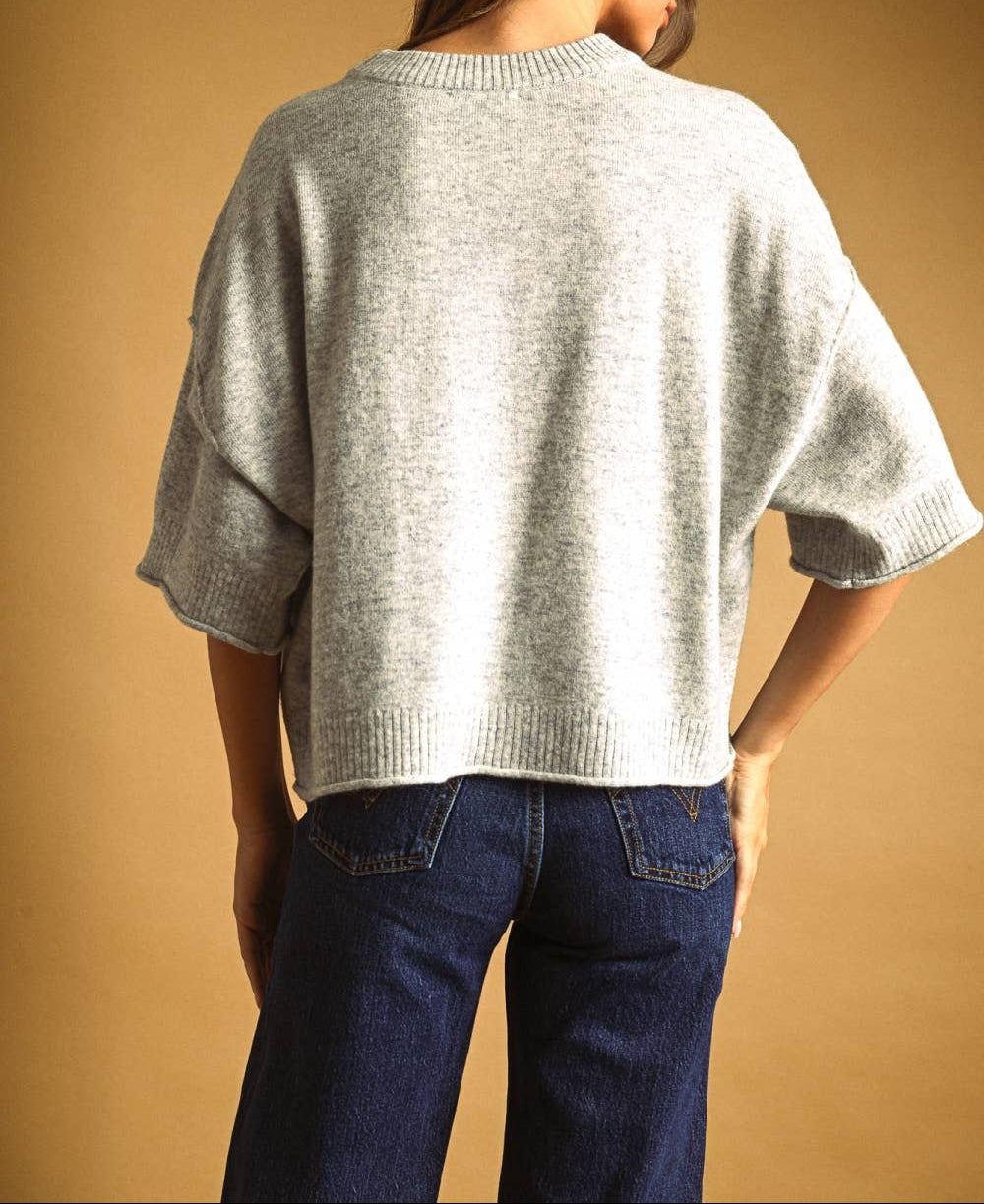 A woman shown from the back wearing a light gray knit sweater with wide three-quarter sleeves and dark blue jeans, set against a solid warm tan background.