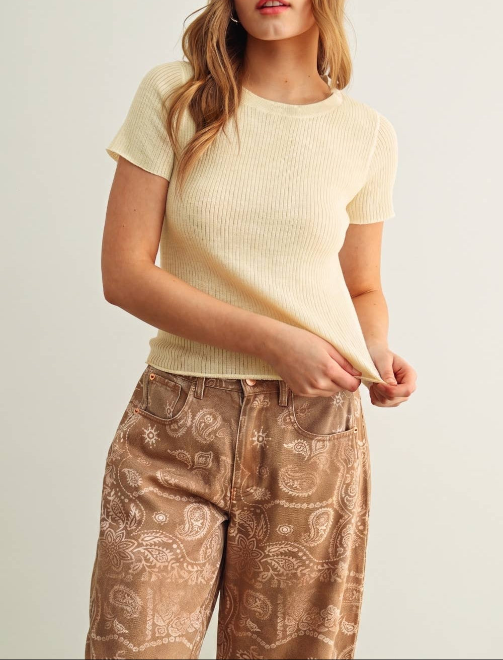 Woman wearing a light yellow ribbed sweater and patterned brown pants on a plain background