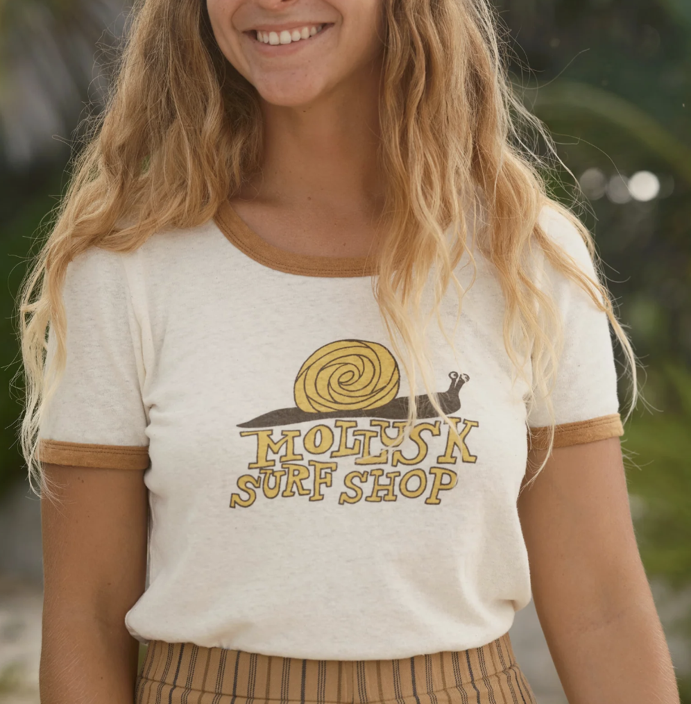 A woman wearing a beige and tan colored t-shirt with 'Mollusk Surf Shop' graphic and a snail illustration.