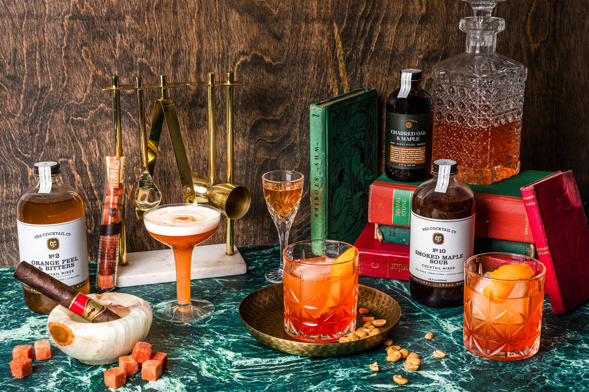 A styled bar scene with several amber cocktails in glasses, bottles of orange-flavored spirit, a decanter, mixing tools, books, and a marble surface against a dark wood backdrop.