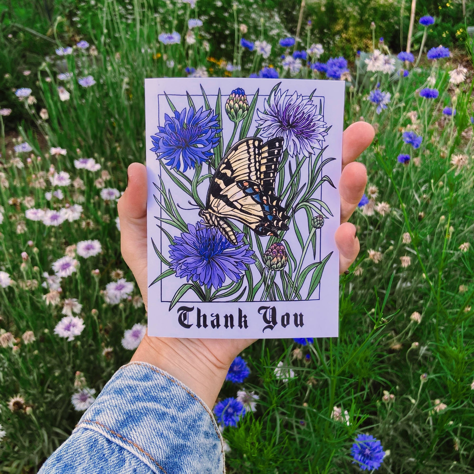 Hand holding a card with a butterfly and flowers design in front of a garden background