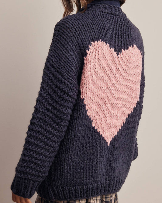 A woman shown from the back wearing a chunky navy blue knit cardigan with a large pink heart on the back, paired with a plaid mini skirt and a beige beret, against a plain light gray studio background.