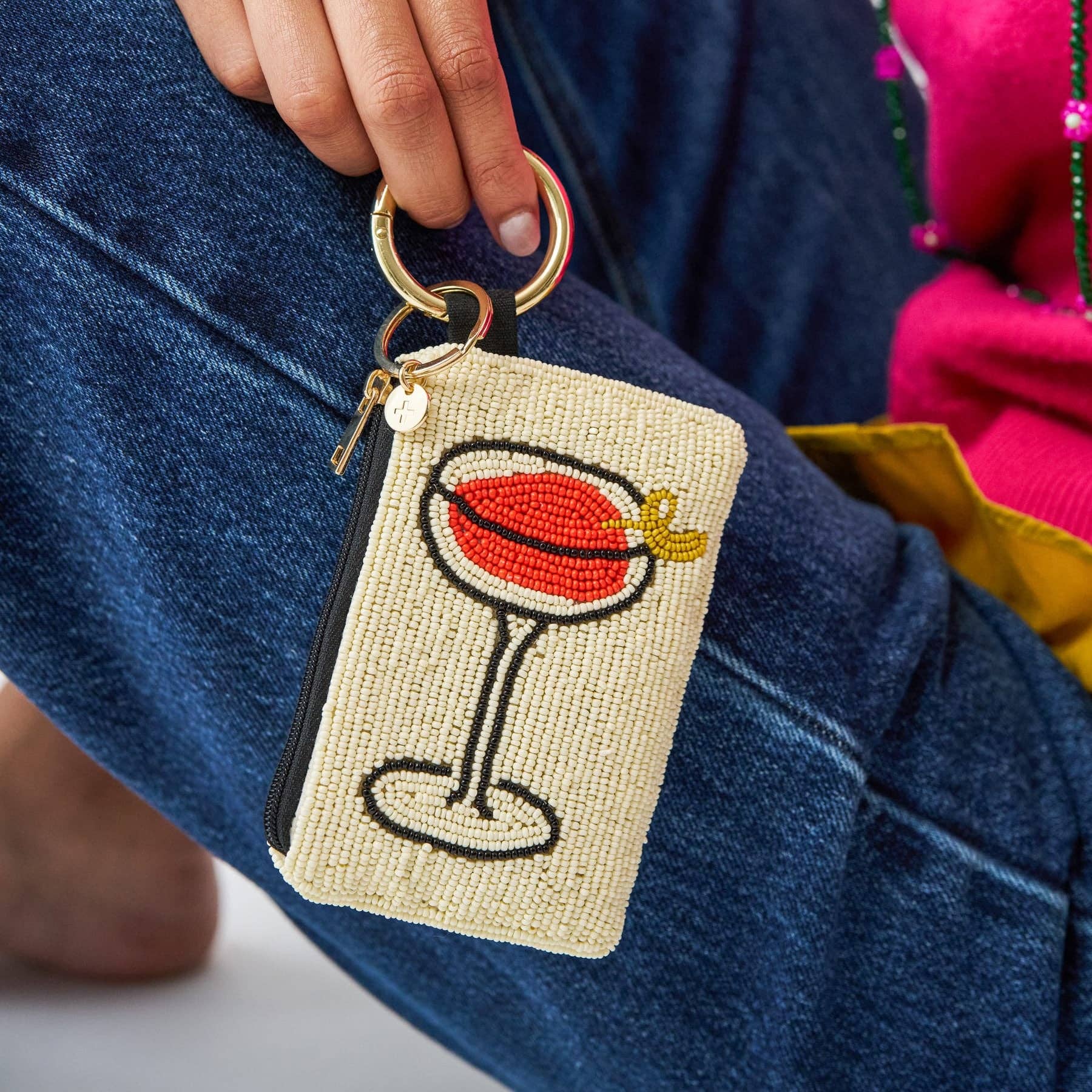 A close-up of a person wearing blue denim jeans, holding a small woven pouch with a gold key ring. The pouch is beige with a beaded cocktail glass design featuring red, black, and yellow details.