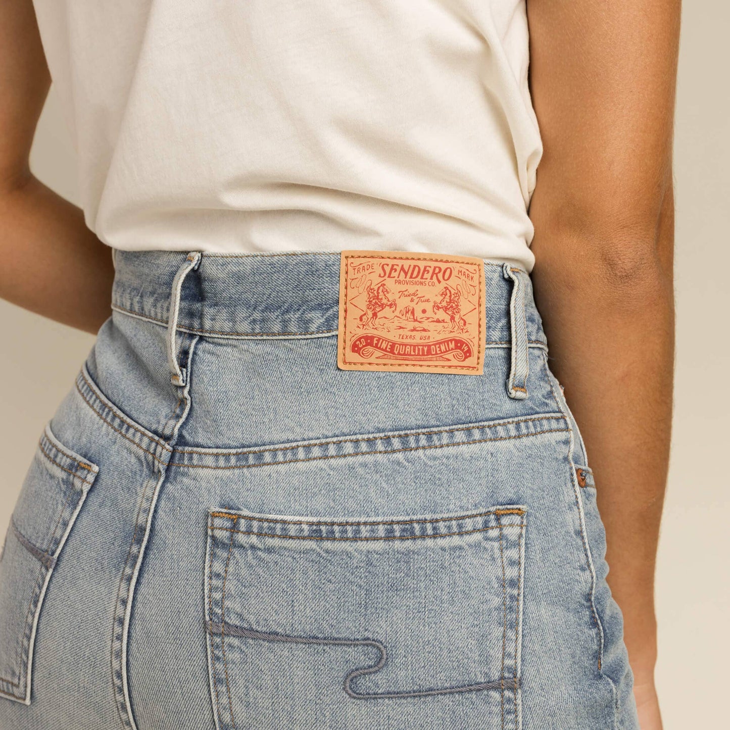 Close-up of a person wearing blue jeans with a Sendero label on a neutral background
