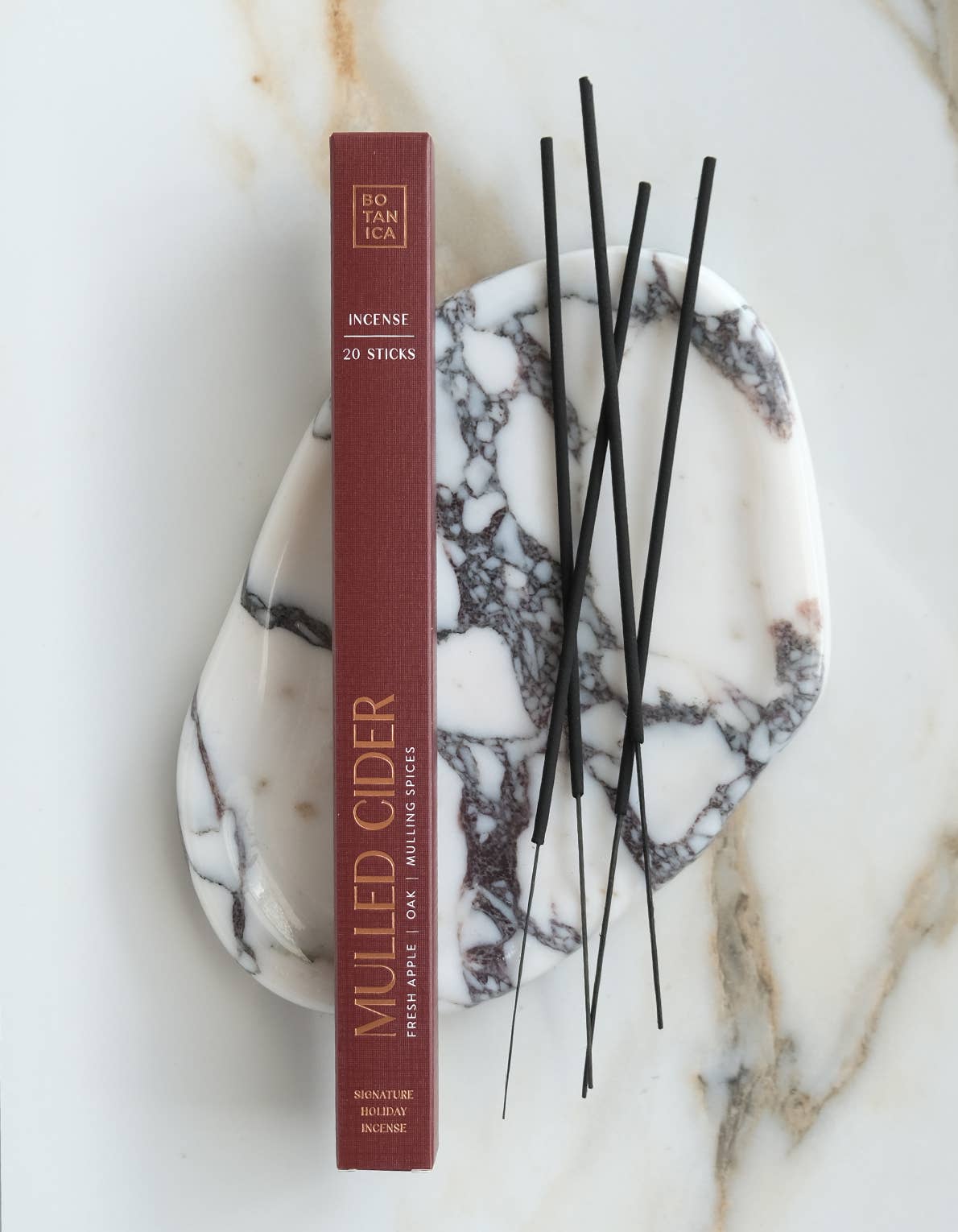 A vertical box of incense labeled 'MULLED CIDER' stands on a white marble tray with black incense sticks, photographed on a light marble surface.