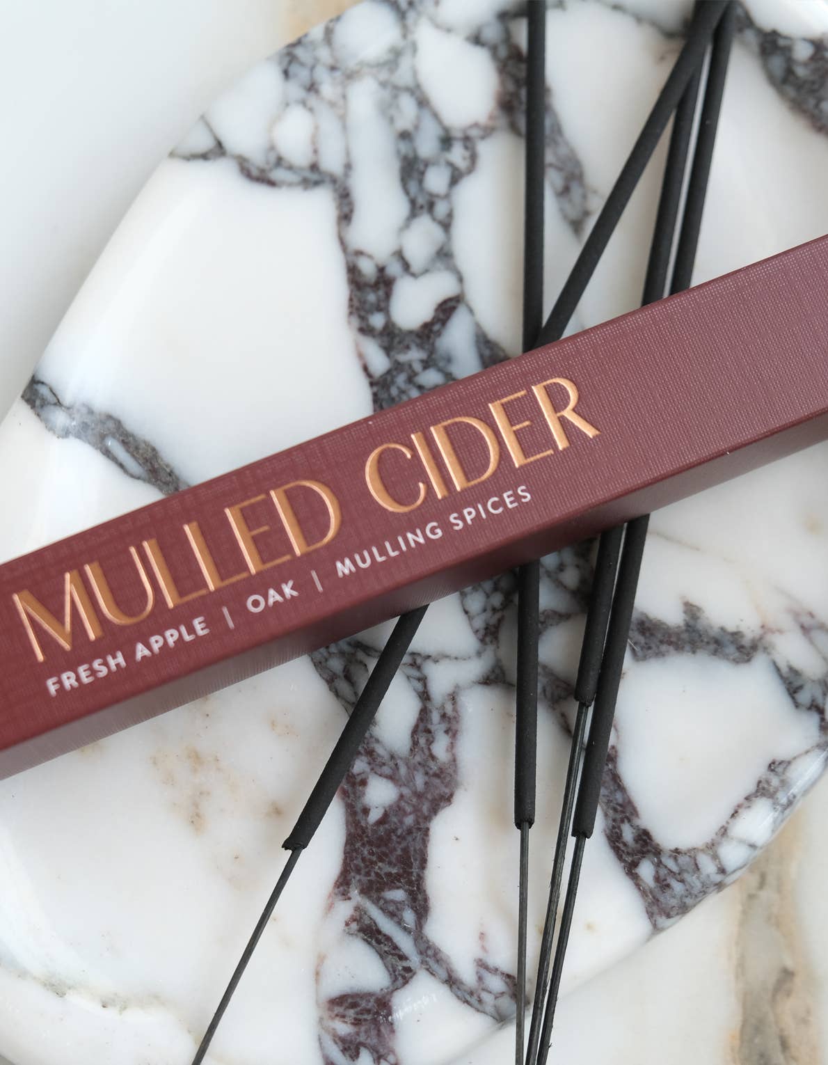 A close-up of a spice box labeled 'MULLED CIDER' with gold lettering on a dusty burgundy-red box, placed diagonally on a white and gray marble surface with several black incense sticks.