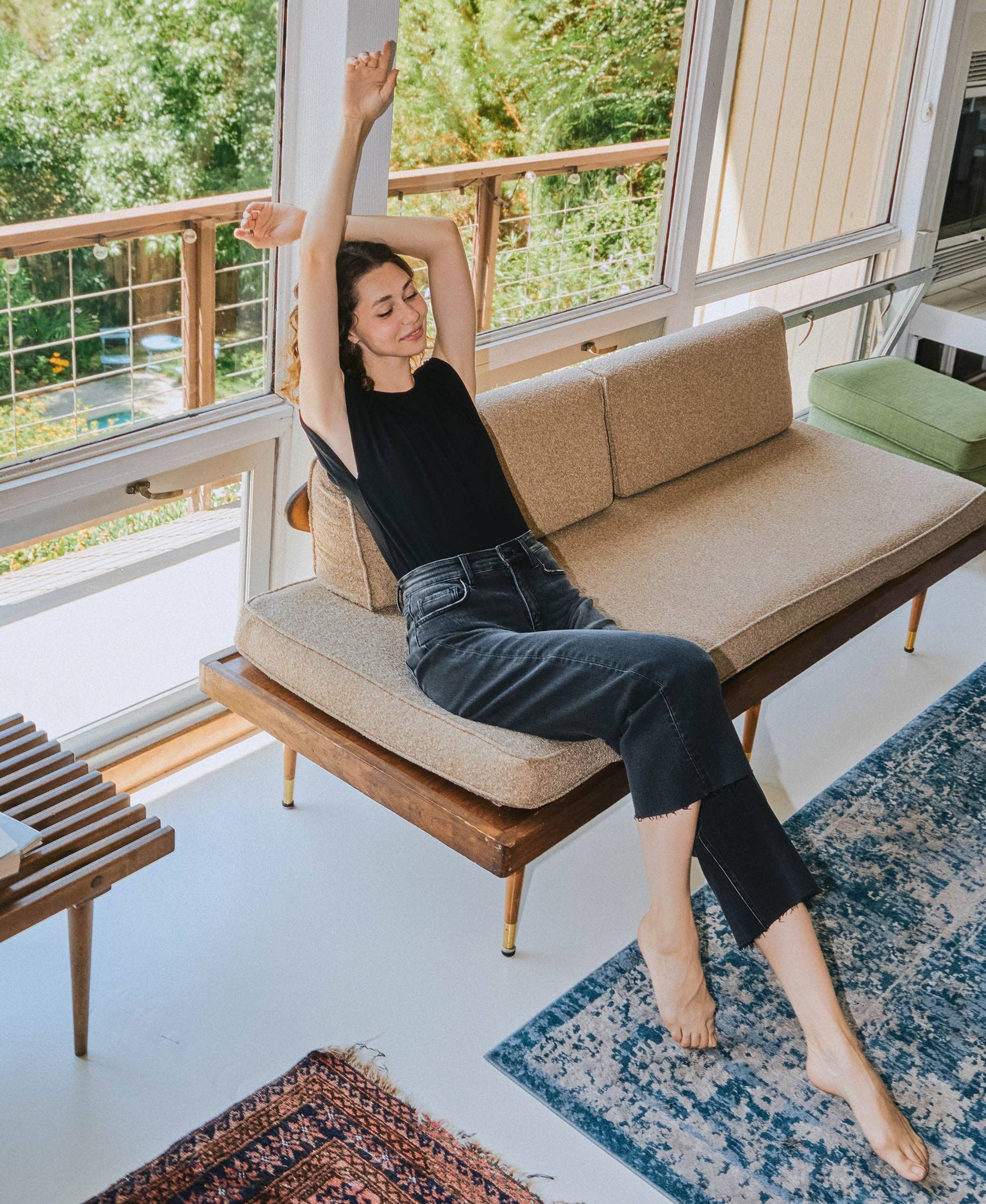 Woman stretching on a modern sofa in a bright living room with large windows.