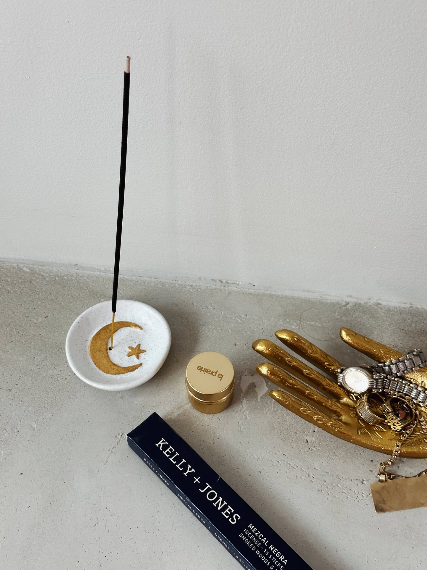 Incense stick with a decorative dish on a concrete surface, next to a gold hand sculpture and a Kelly & Jones product.