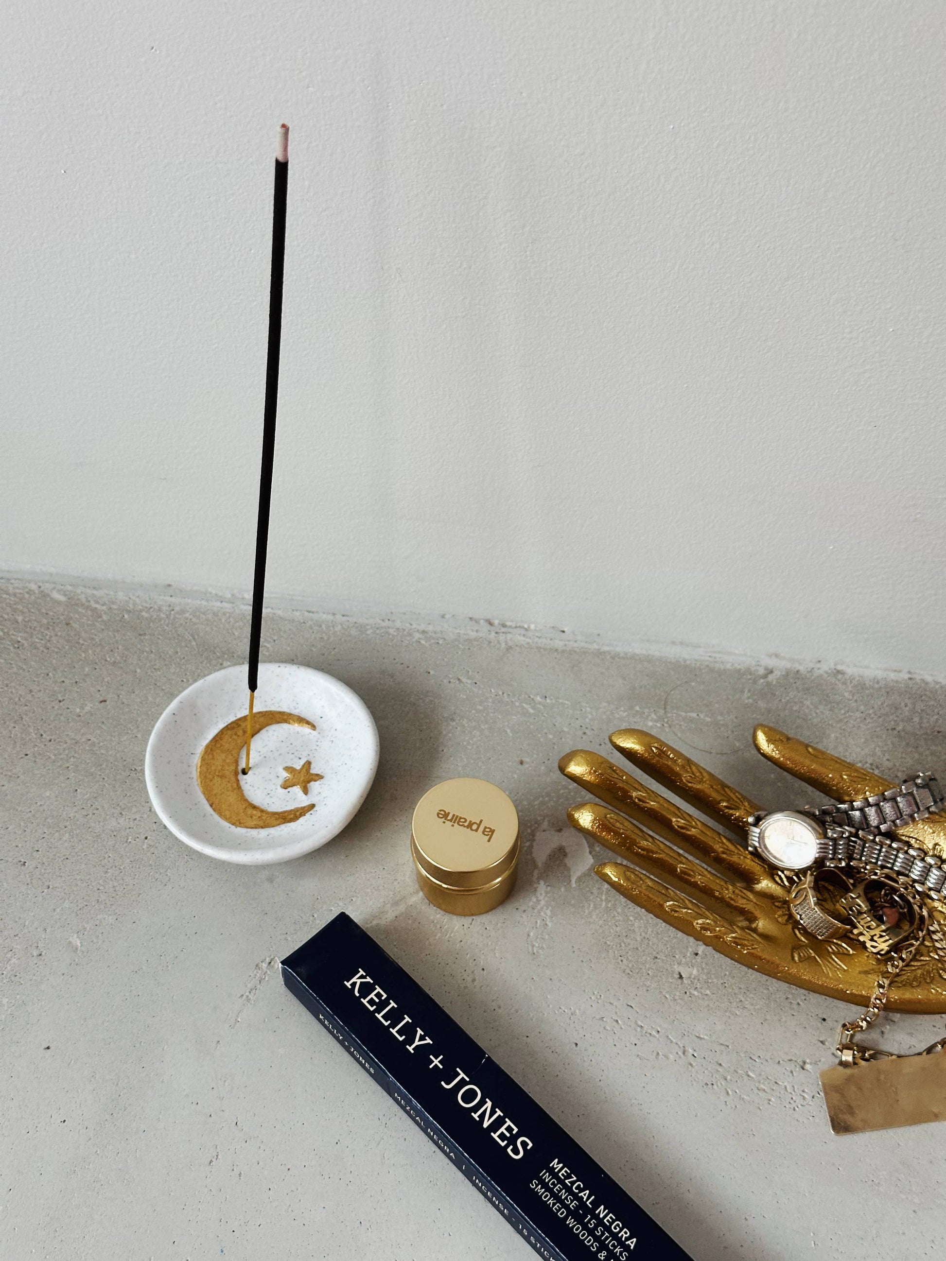 Incense stick with a decorative dish on a concrete surface, next to a gold hand sculpture and a Kelly & Jones product.